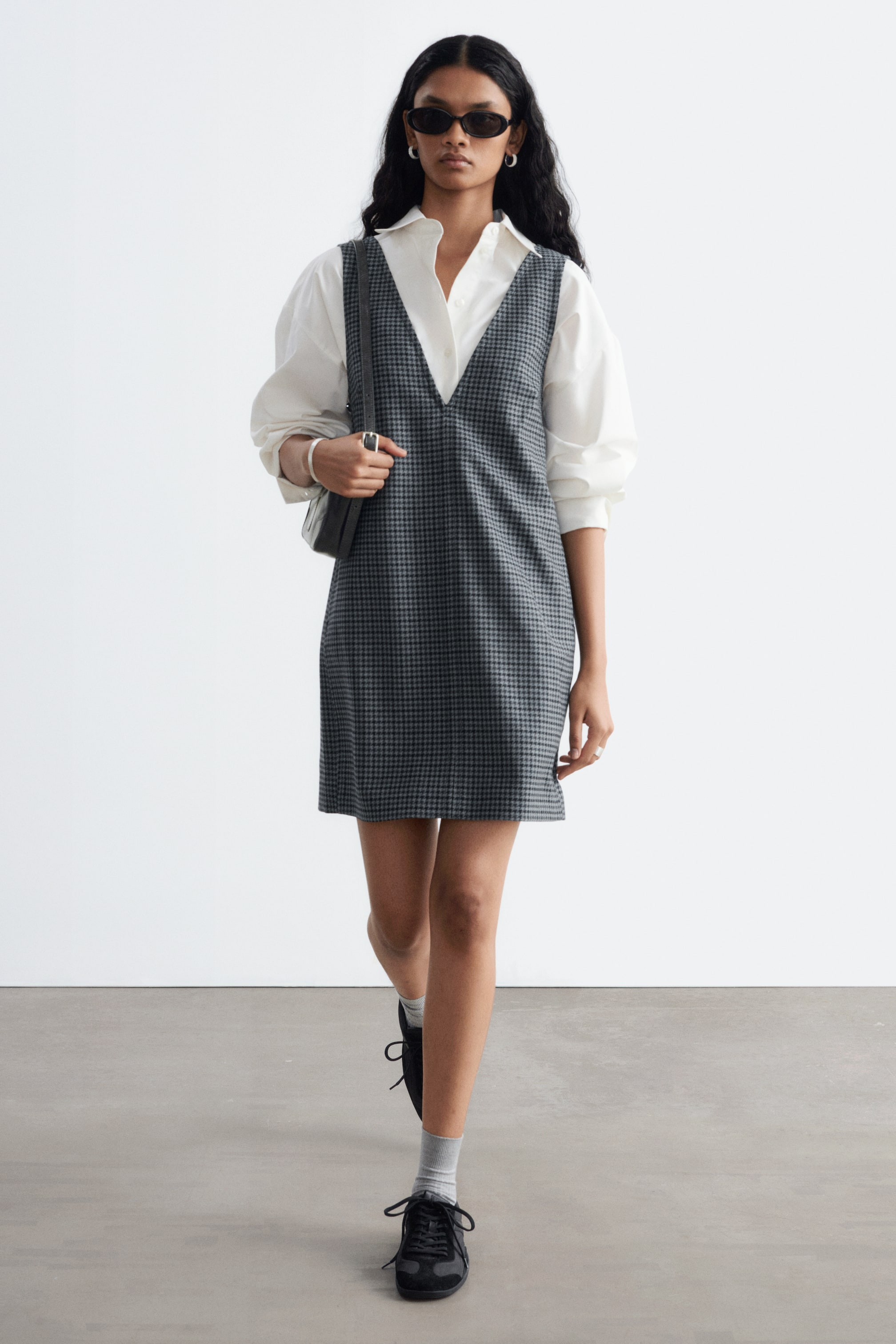 Ver imagen más grande: La modelo lleva un vestido pichi de pata de gallo en gris y negro con un pronunciado escote en V, superpuesto sobre una camisa blanca de manga larga y cuello. El vestido sin mangas y de corte recto se combina con un bolso negro de hombro llevado a un lado.