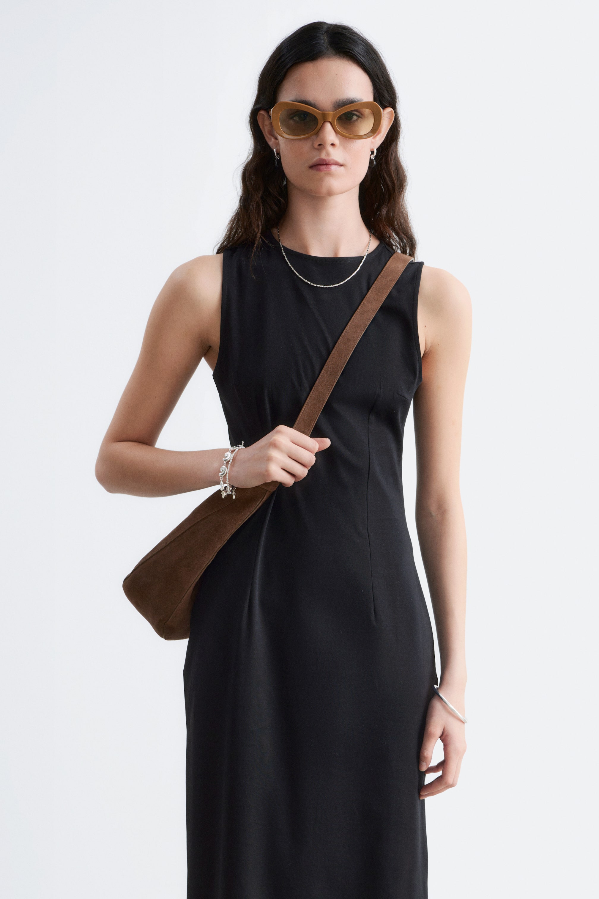 Agrandir l'image: La modèle porte une robe midi noire sans manches avec un col rond et une silhouette ajustée. Un sac à bandoulière marron est porté par-dessus la robe.