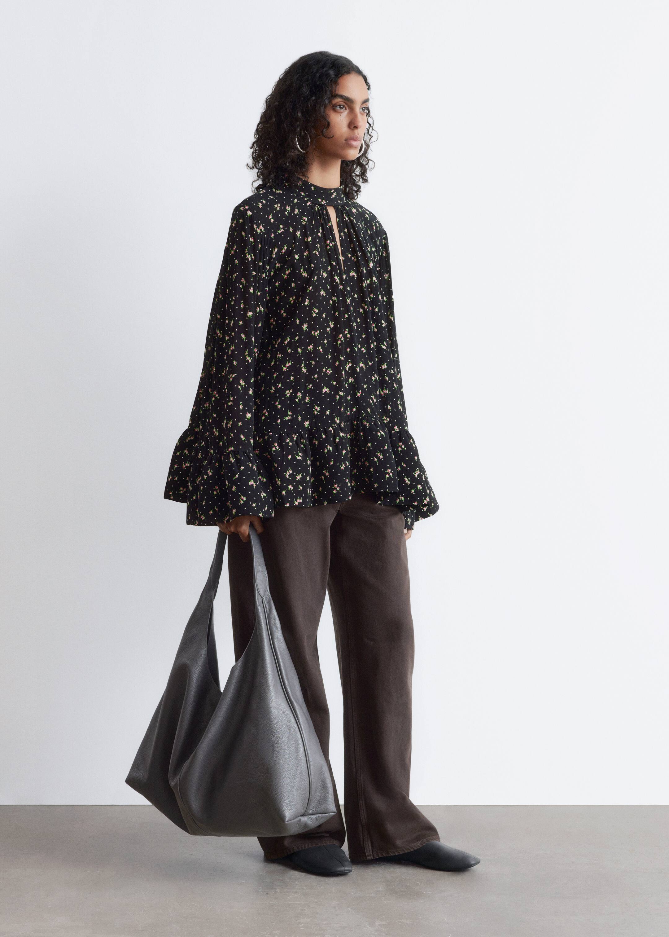 Ver imagem maior: Uma blusa preta com uma pequena estampagem floral e de bolinhas está a ser apresentada de um ângulo frontal-direito, apresentando um colarinho alto com uma abertura em forma de gota e um detalhe de laço. Está a mostrar mangas compridas e largas com punhos franzidos e uma silhueta solta e fluida com uma bainha em camadas franzida.