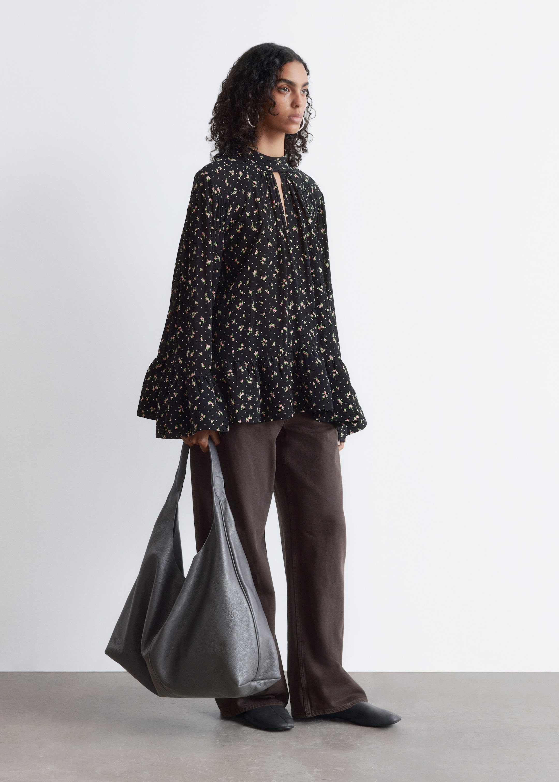 Agrandir l'image: Une blouse noire avec un petit imprimé floral et à pois est présentée sous un angle avant-droit, dotée d'un col montant avec une ouverture en goutte d'eau et un détail de nœud. Elle présente de longues manches larges avec des poignets à volants et une silhouette ample et fluide avec un volant froncé à l'ourlet.