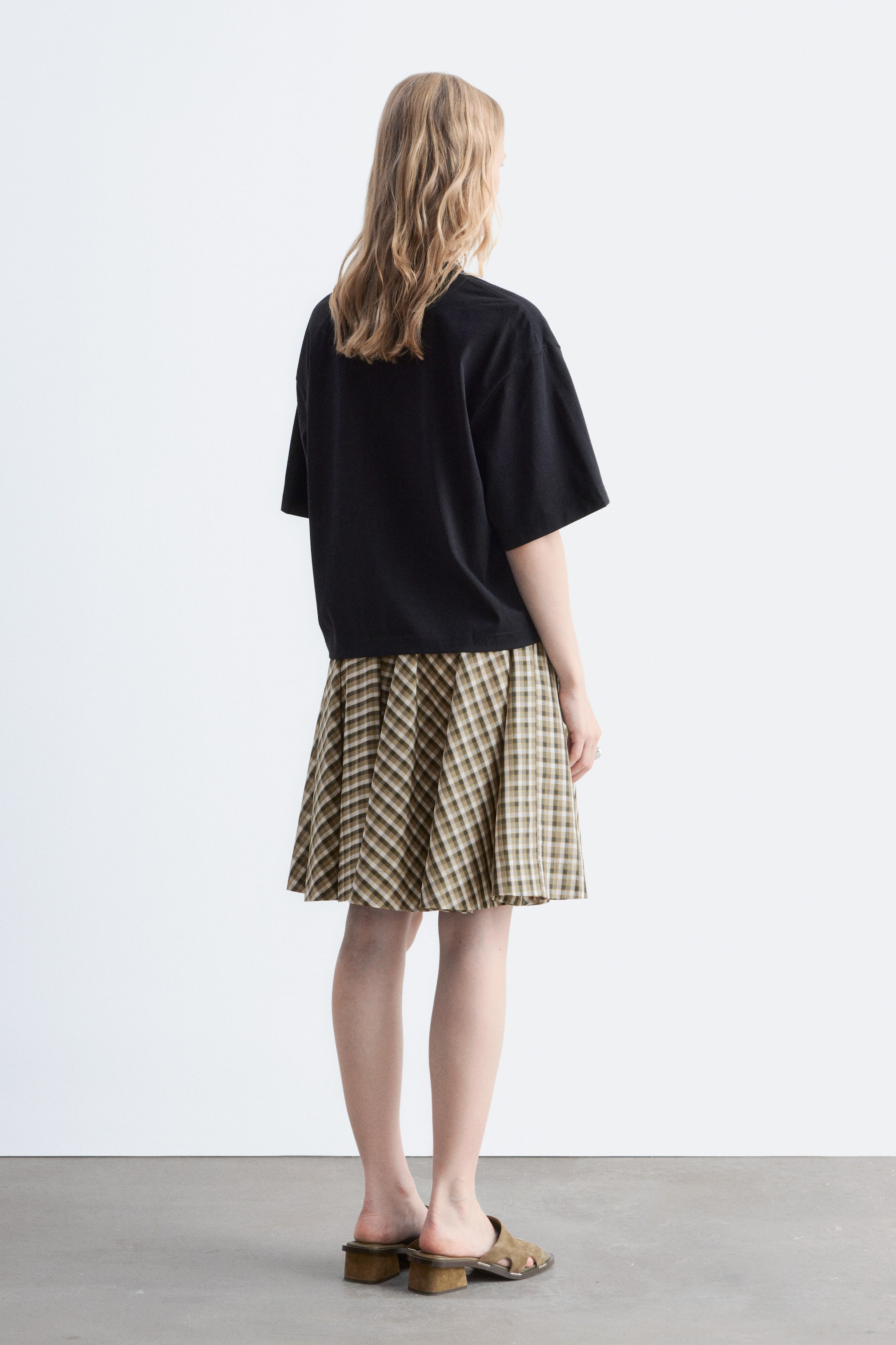 Grotere afbeelding bekijken: Een zwart T-shirt, met een ontspannen, boxy vorm en korte, brede mouwen, wordt gedragen door het model. Het T-shirt is gestyled met een geruite rok.