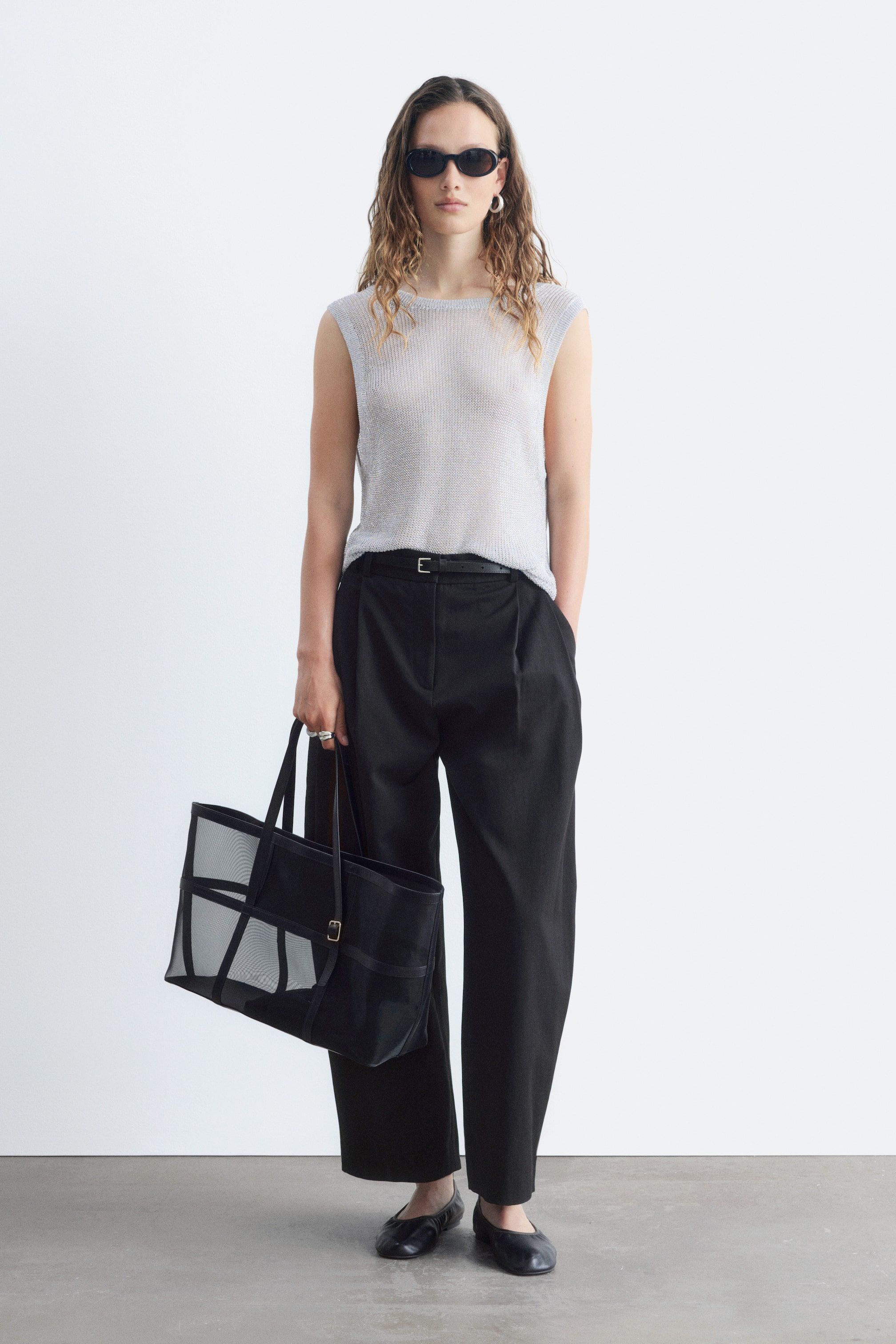 View larger image: An ensemble featuring a shimmering silver sleeveless top with a boat neckline, black pleated wide-leg trousers cinched by a thin black belt, and black flat shoes. A black mesh tote bag with a grid panel design is being held, presenting the complete outfit from a front-facing angle.