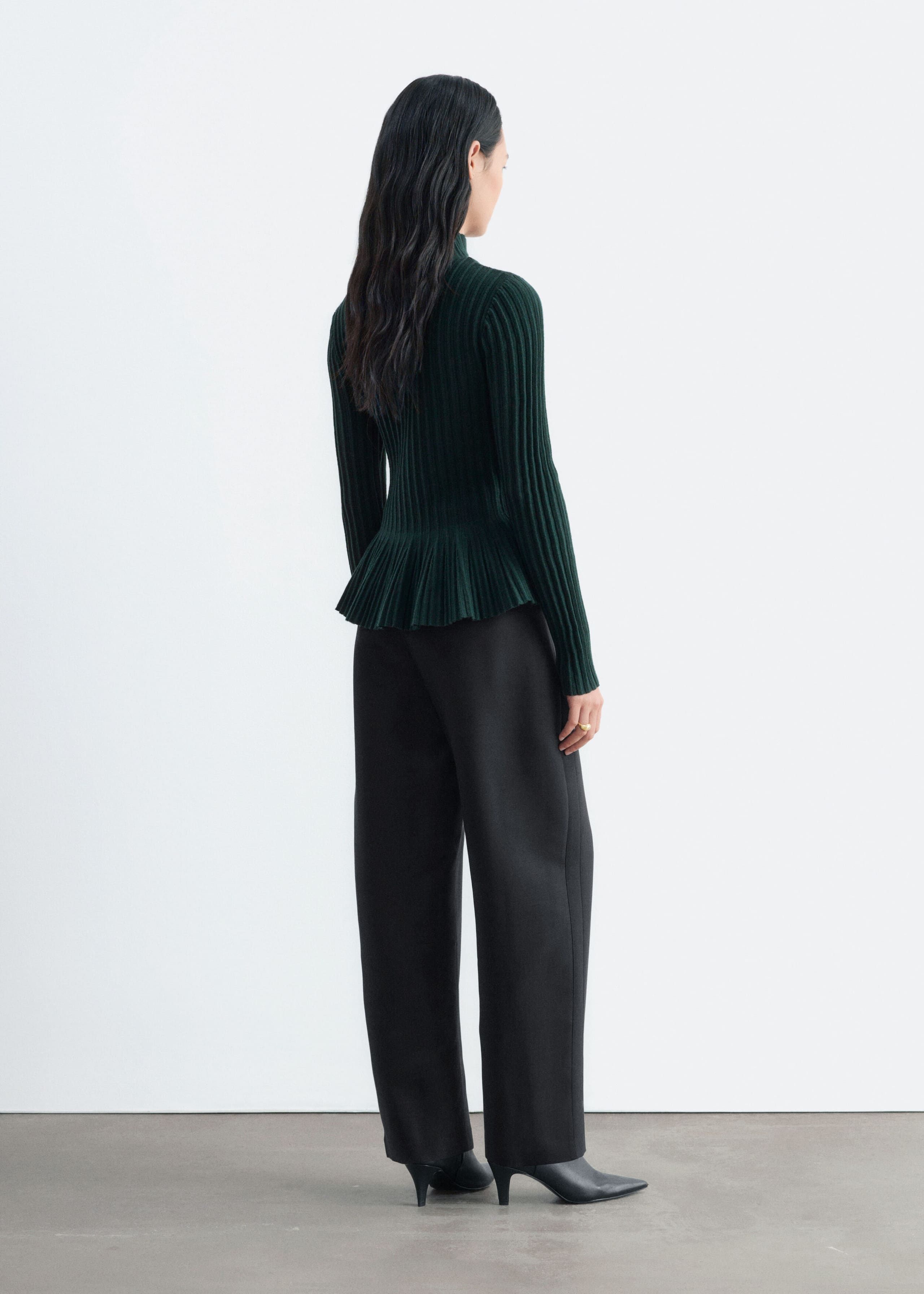 The model is wearing a dark green ribbed-knit top featuring a mock neck, long sleeves, and a flared peplum hem. The top is styled with wide-leg black trousers and black pointed-toe heeled shoes.