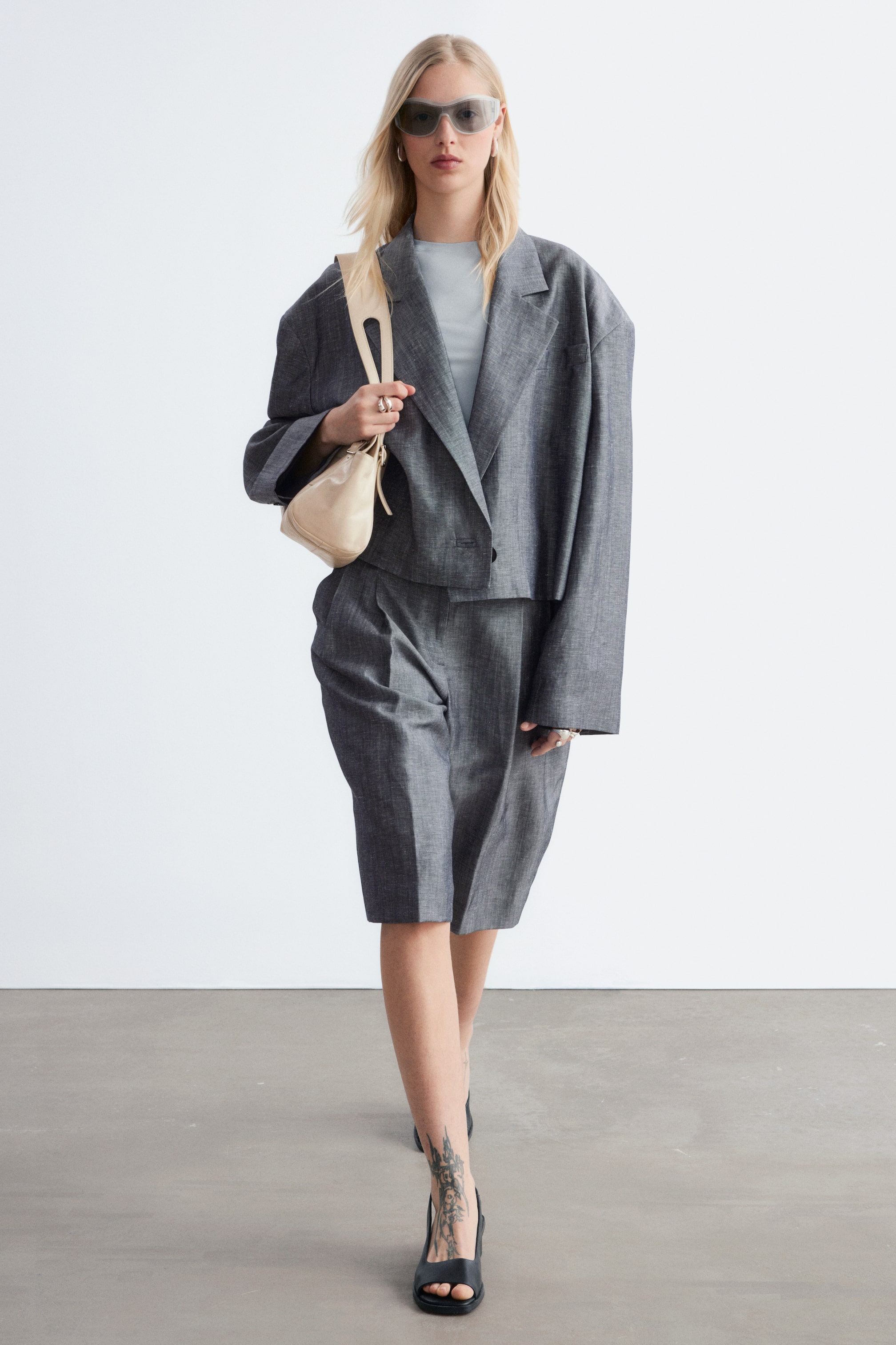 Ver imagem maior: Conjunto de fato cinzento texturizado em duas peças, composto por um blazer oversized e cropped com lapelas em bico e mangas largas, usado aberto sobre uma blusa azul clara. Calções Bermuda a condizer, com pregas frontais e bolsos laterais, completam o conjunto, estendendo-se até meio da coxa.