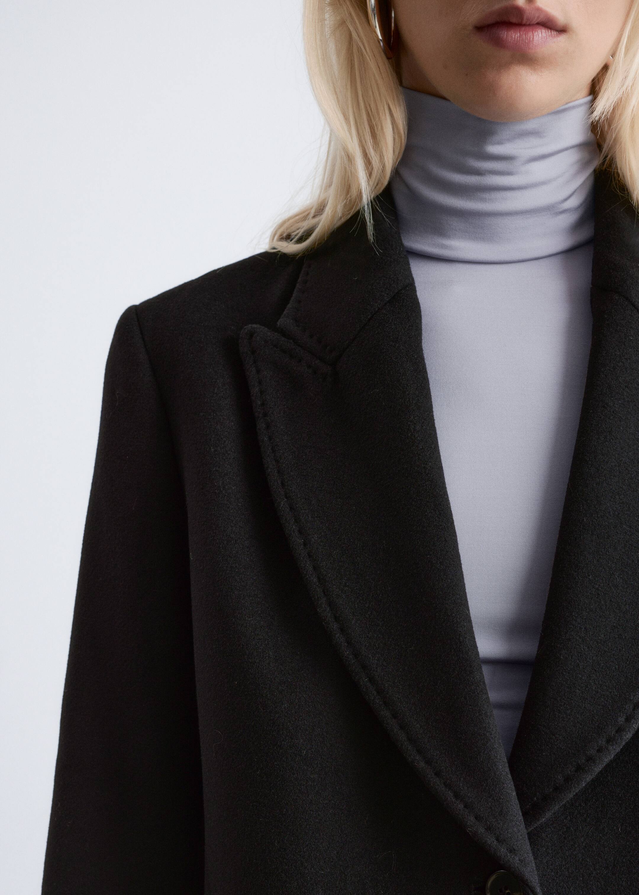 Agrandir l'image: Un manteau noir est doté d'épaules structurées et de revers en pointe avec une couture visible sur les bords, présenté en angle de face. Il est superposé à un col roulé gris clair, et un bouton unique est visible près du bas.
