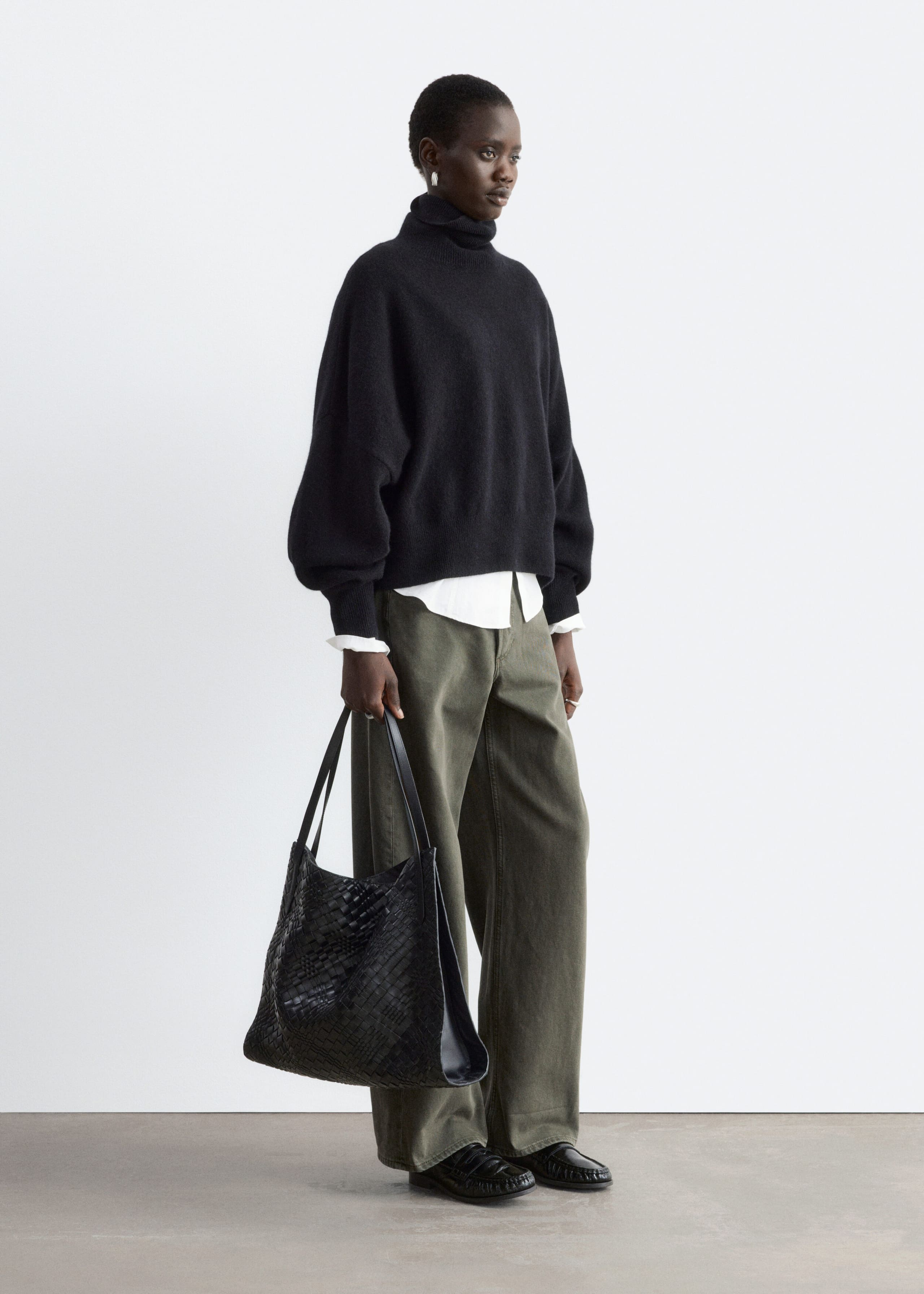 An oversized black roll neck jumper with dropped shoulders and ribbed cuffs is being worn by the model over a layered white shirt. The outfit is completed with green wide-leg trousers and a black woven tote bag being carried by the model.