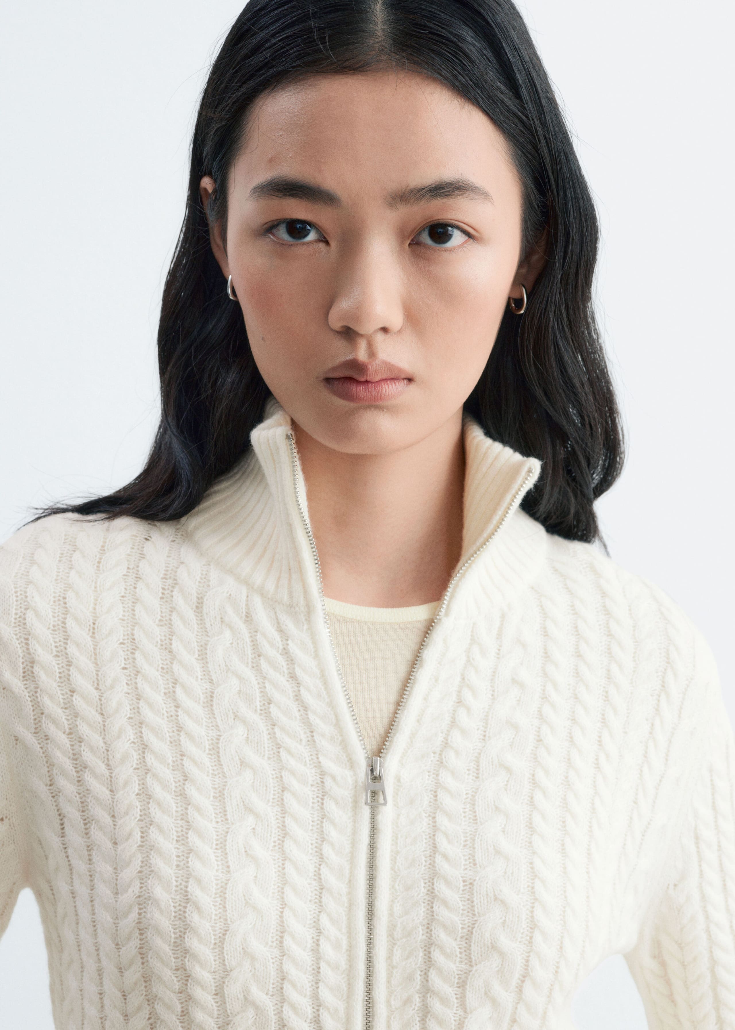 The model is wearing a cream cable-knit cardigan, featuring a partially open silver zip and a high ribbed collar. The cardigan is layered over a light cream top.