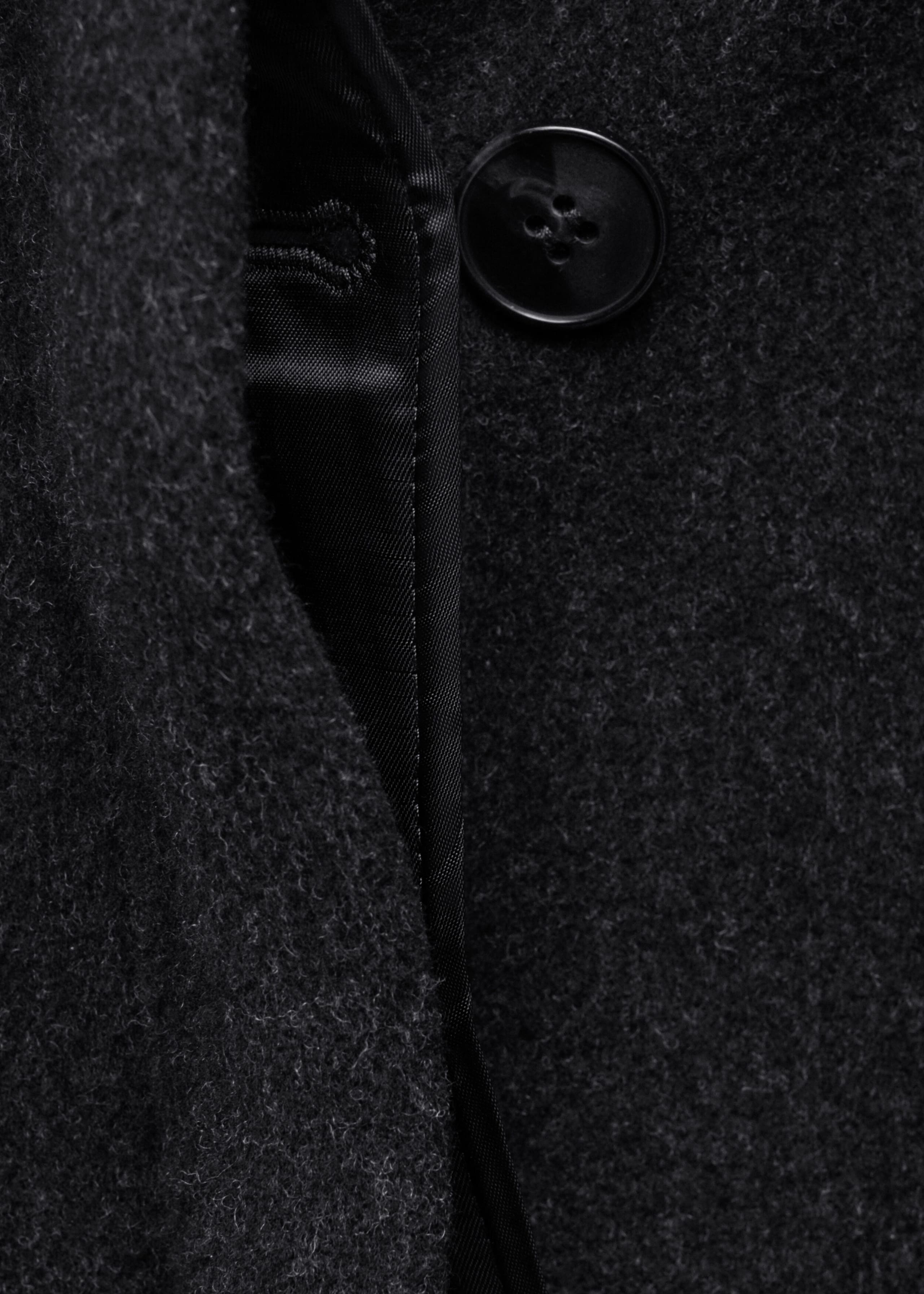 A dark charcoal wool-blend coat is shown in a close-up view, highlighting its textured, felted fabric and interior construction. The image reveals a smooth black satin lining, a stitched buttonhole, and a black four-hole button forming the concealed front fastening.