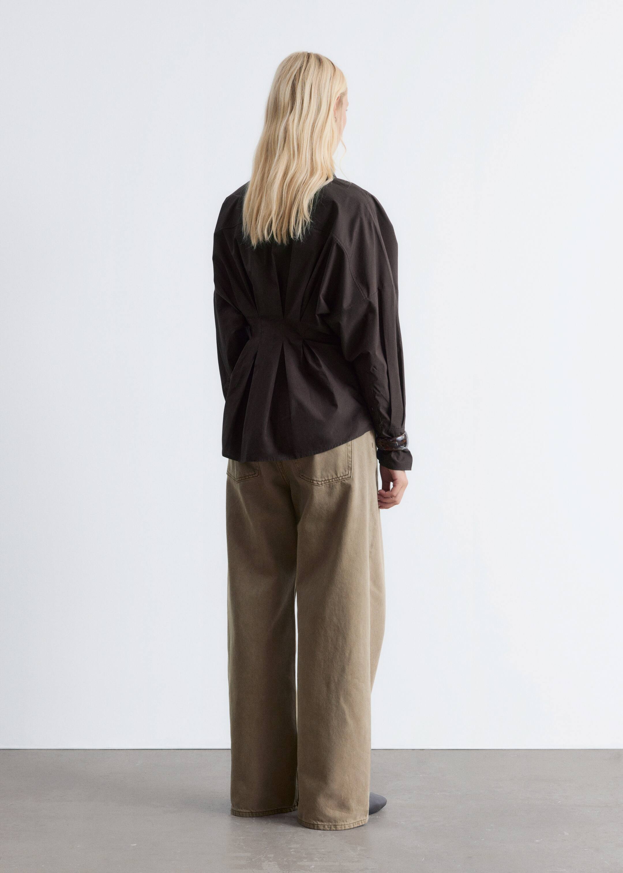 View larger image: A dark, long-sleeved shirt is being presented from the back, featuring a cinched waistline and prominent pleats below it that add volume. It is being styled with light brown, wide-leg pants that have a relaxed fit.