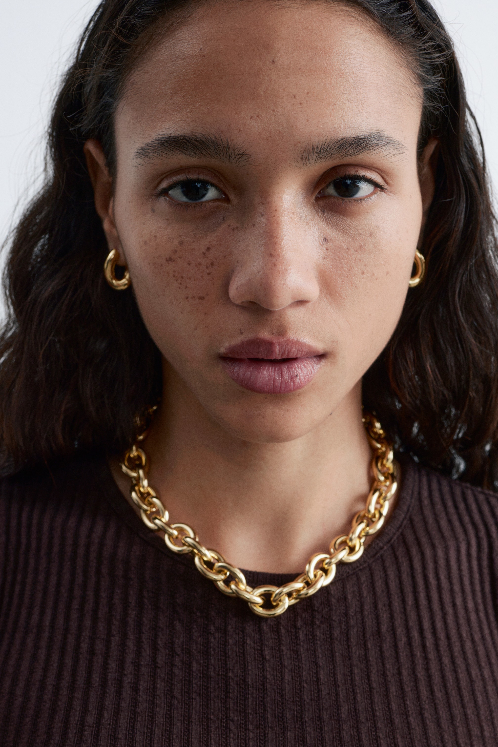 Ver imagen más grande: Un collar de cadena gruesa en tono dorado descansa sobre un top acanalado marrón, con grandes eslabones ovalados interconectados. Aros gruesos a juego en tono dorado se llevan en cada lóbulo de la oreja.