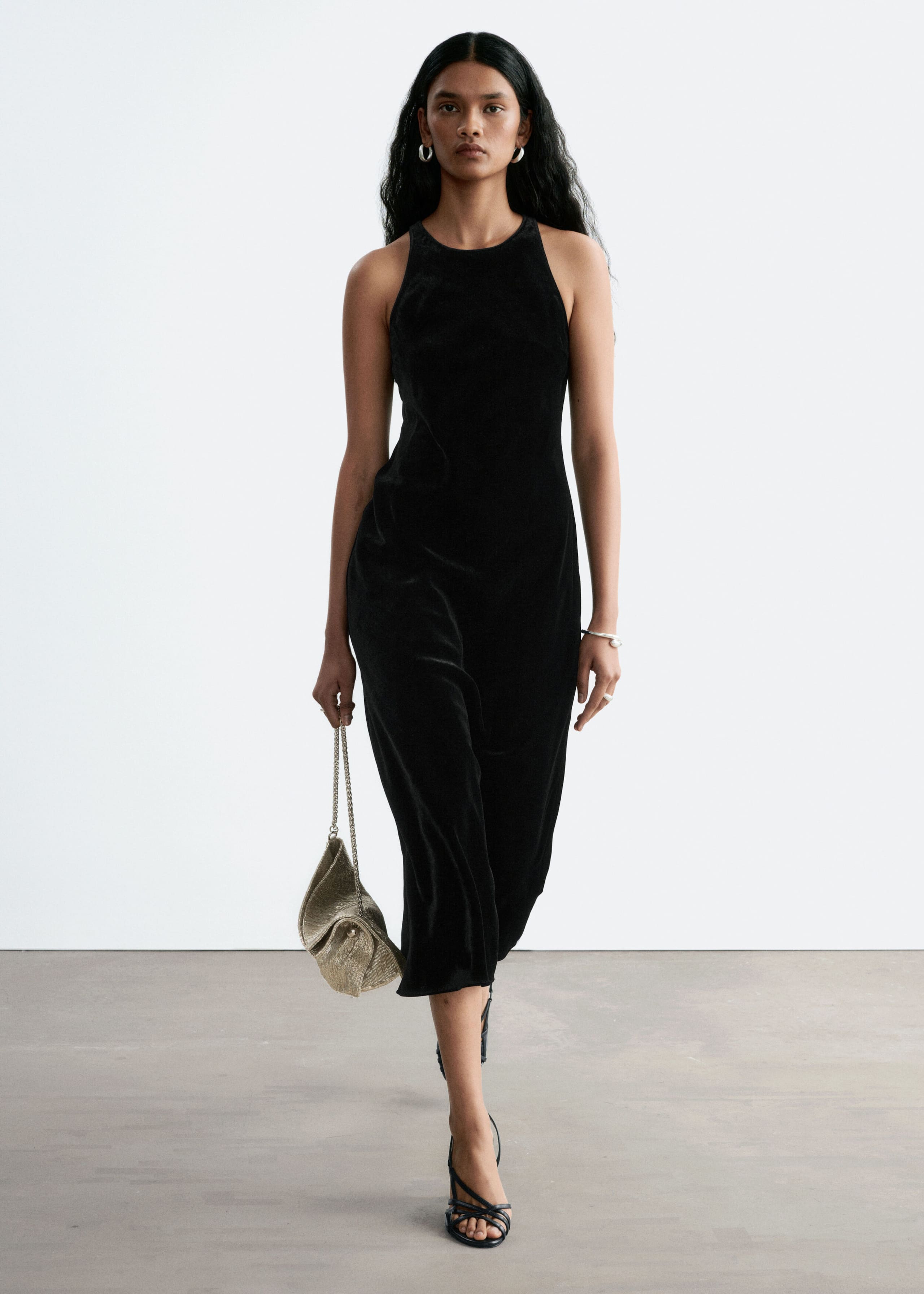 A black, sleeveless midi dress with a high crew neckline and a glossy finish is worn by the model as she walks forward. The dress is styled with black strappy heeled sandals and a small gold-tone handbag held in one hand.