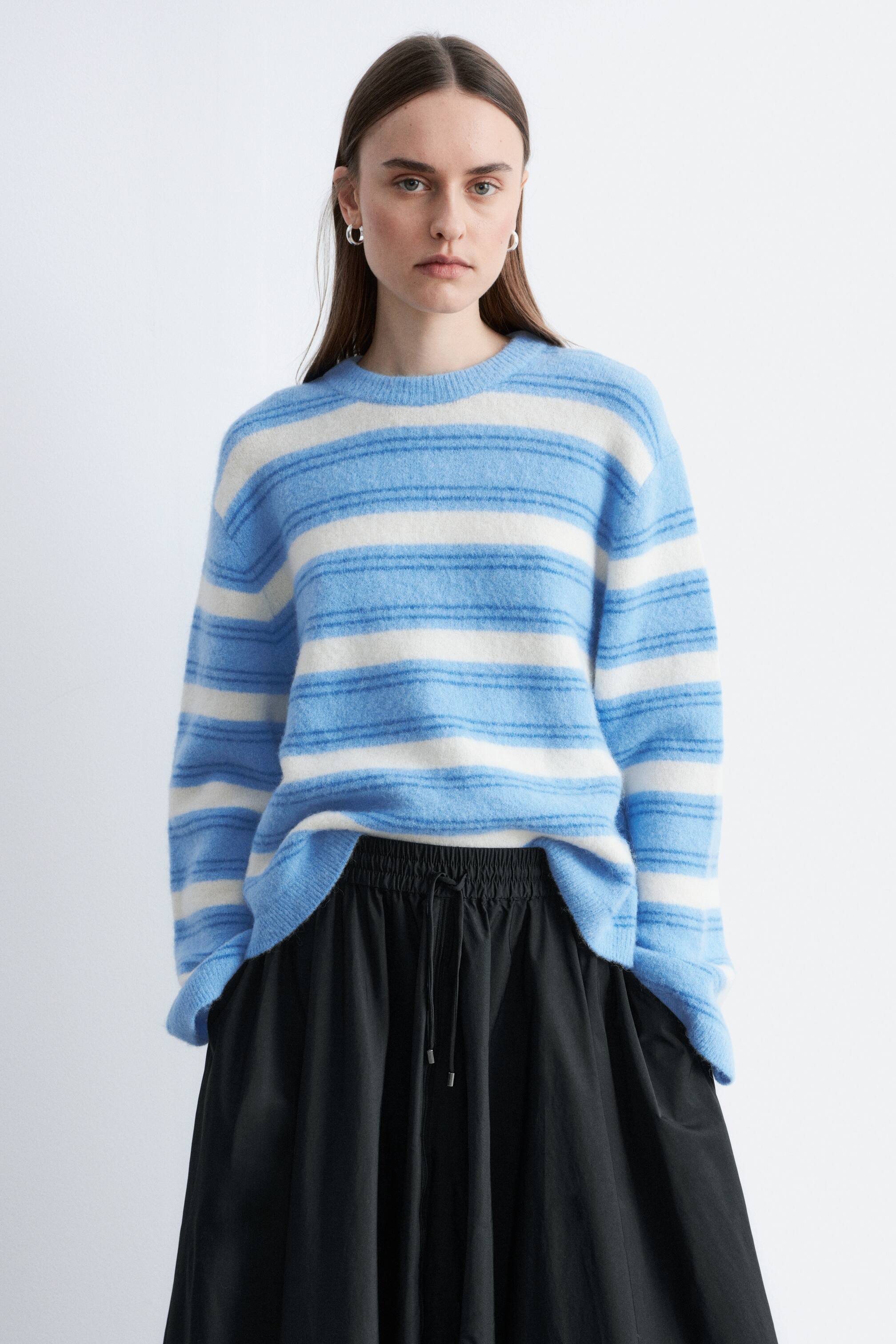 Преглед на по-голямо изображение: Синьо-бялото райе на пуловера подчертава свободен силует, кръгло деколте, спуснати рамене и рипсени кантове. Моделът носи пуловера, който е частично вкаран в черна пола.