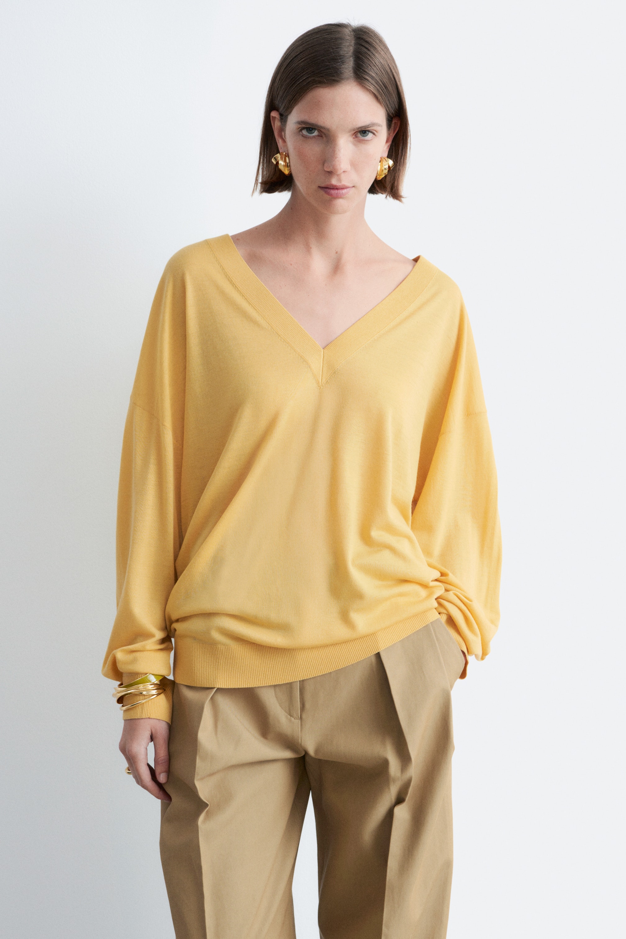 Visualizza immagine più grande: Il maglione giallo con scollo a V presenta una vestibilità rilassata con scollo a V a coste, maniche lunghe e ampie, e un orlo a coste, abbinato a pantaloni color cachi.