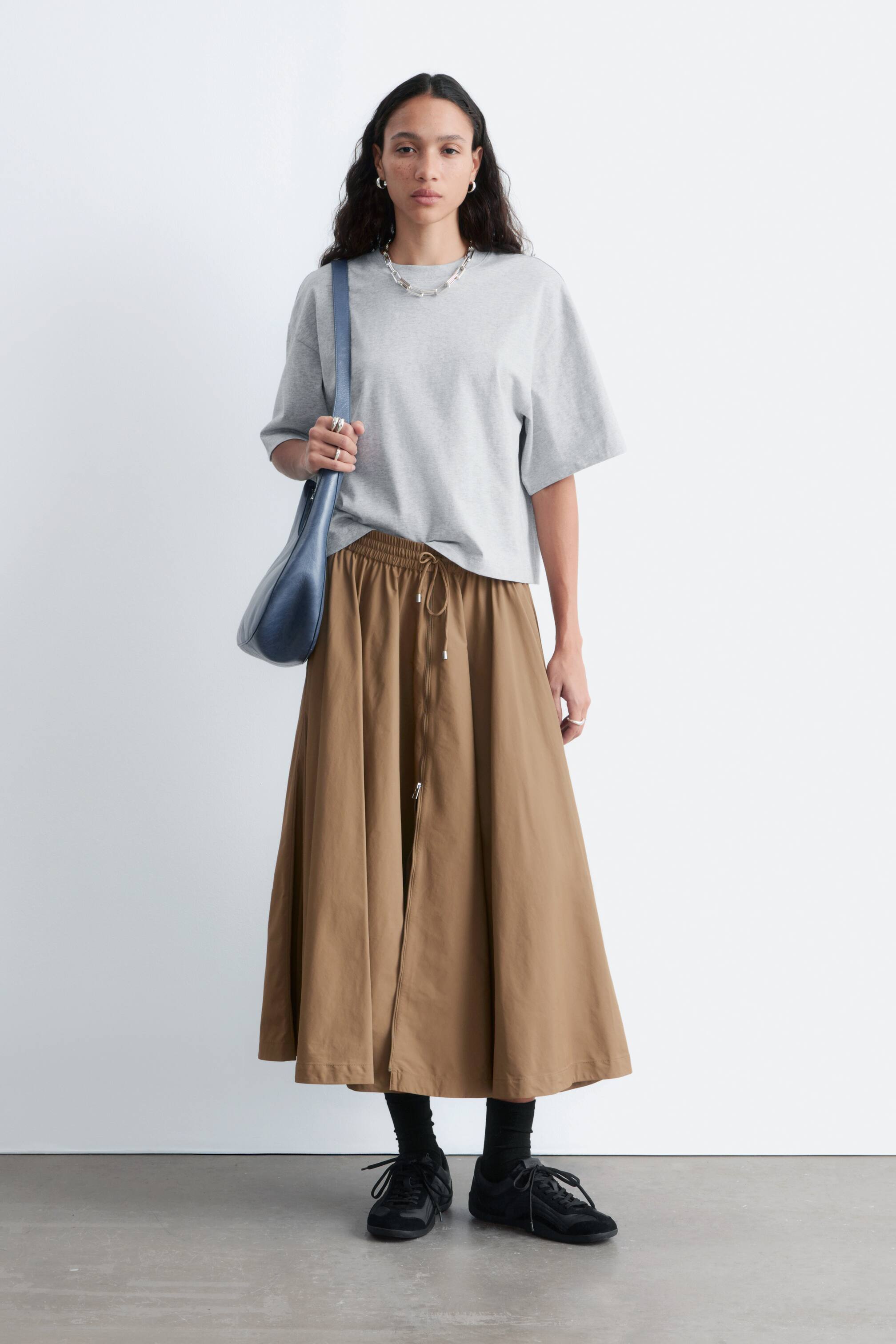 Größeres Bild anzeigen: Der braune Midi-Rock präsentiert sich in einer A-Linien-Silhouette mit einem gerafften, elastischen Kordelzugbund, der von vorne sichtbar ist. Er verfügt über eine zentrale Frontleiste mit Reißverschluss und einem sichtbaren Kordelzug, der in der Taille gebunden ist, wobei der Stoff sanft zu einer Midi-Länge fließt.