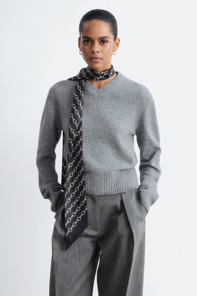 La modelo lleva un jersey de punto gris con escote en V acanalado, bajo ancho acanalado y largos puÃ±os acanalados. El jersey se combina con una bufanda de estampado negro y dorado alrededor del cuello y con pantalones grises. - 1