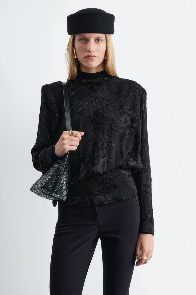 Eine schwarze Chiffonbluse mit floraler Struktur, Mock-Neck und markanten Schulterpolstern wird von dem Model getragen. Die Bluse hat eine geraffte Taille und lange Ãrmel mit Manschetten und wird mit einer schwarzen Hose kombiniert, wÃ¤hrend eine schwarze gewebte Schultertasche Ã¼ber der Schulter getragen wird. - 1
