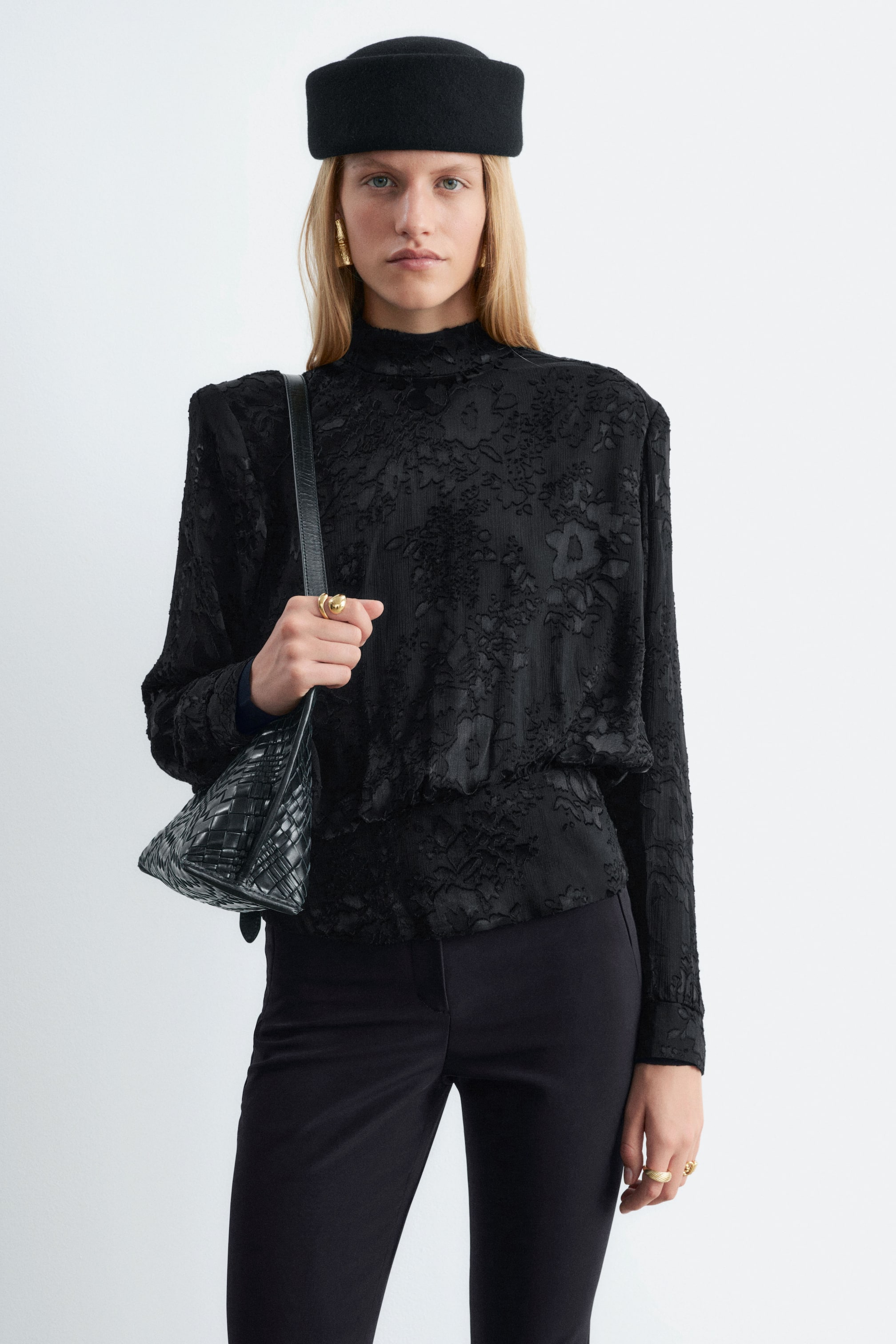 Visualizza l'immagine ingrandita: Una blusa nera in chiffon con texture floreale, caratterizzata da collo alto e spalline strutturate, indossata dalla modella. La blusa presenta un punto vita arricciato e maniche lunghe con polsini, ed è abbinata a pantaloni neri, mentre una borsa a spalla intrecciata nera è portata a tracolla.