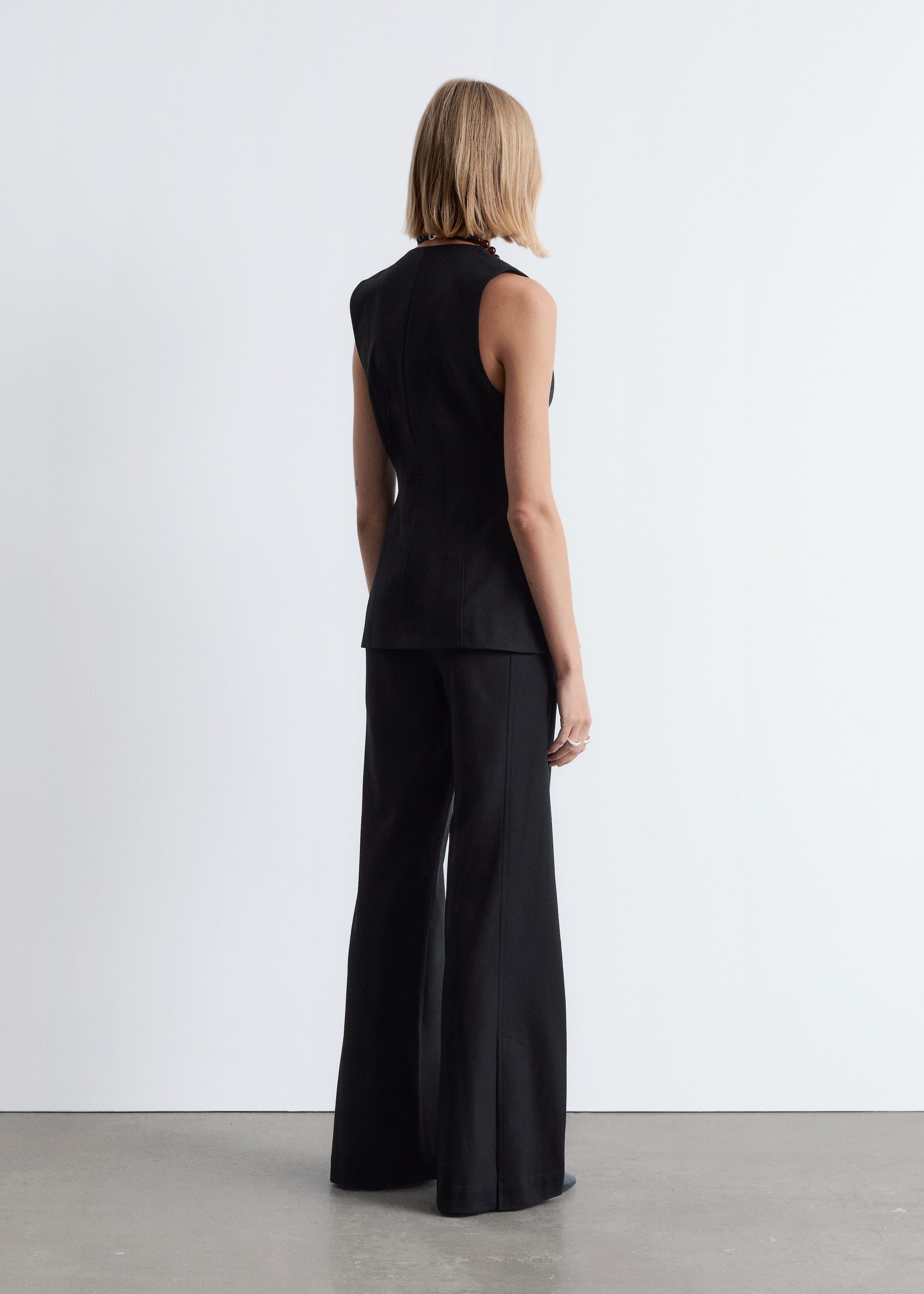 Ingrandisci l'immagine: Un top nero senza maniche e pantaloni neri a gamba larga sono presentati su una figura, vista da dietro e leggermente angolata a destra. Il top è aderente in vita e presenta una cucitura verticale sul retro, mentre i pantaloni hanno una piega stirata che corre lungo la parte anteriore di ciascuna gamba.