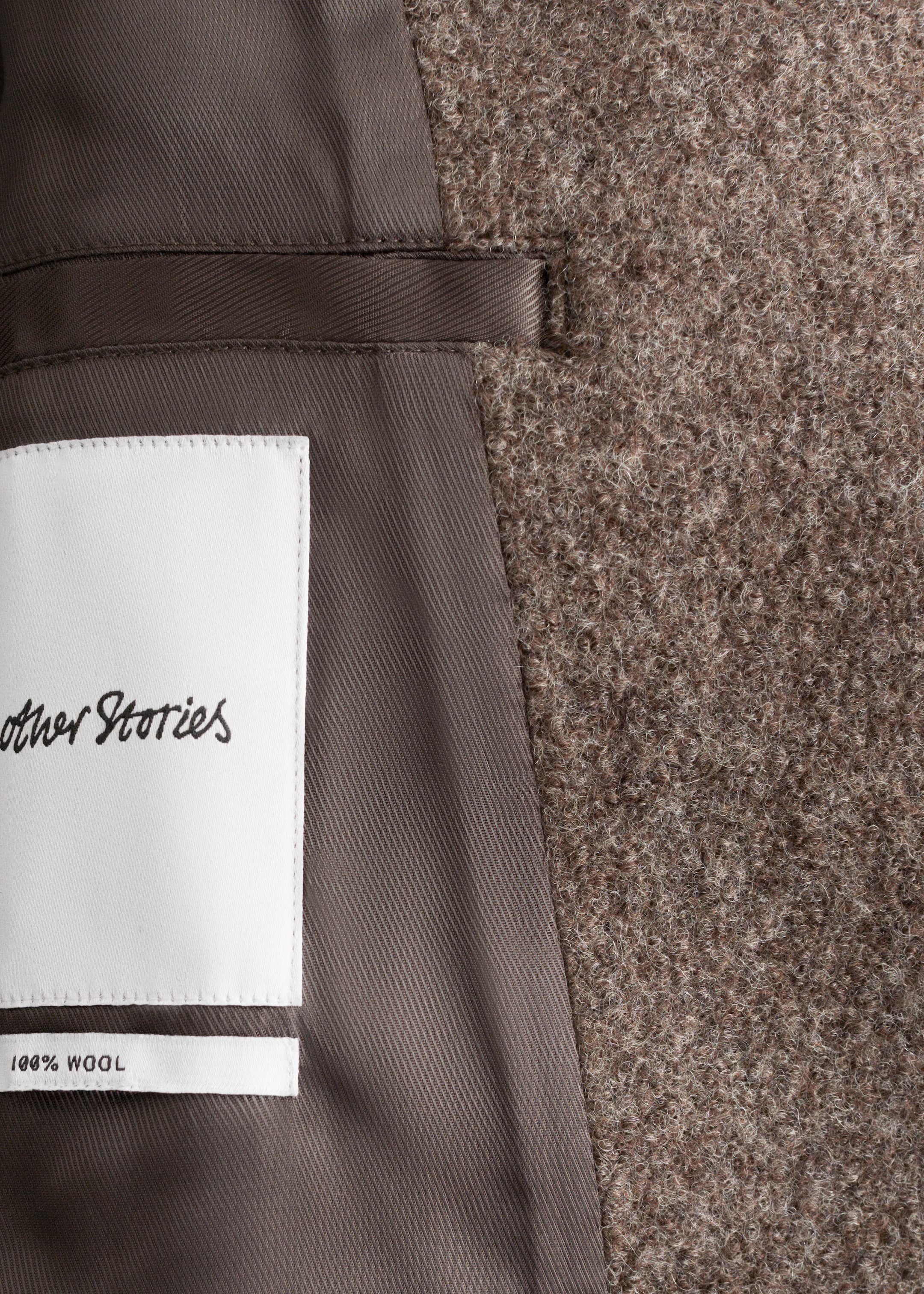 View larger image: A close-up view of the interior of a wool jacket is being shown, revealing its textured brownish-gray outer fabric and smooth dark brown lining. A white brand label, reading 'other Stories' and '100% WOOL,' is stitched onto the lining.
