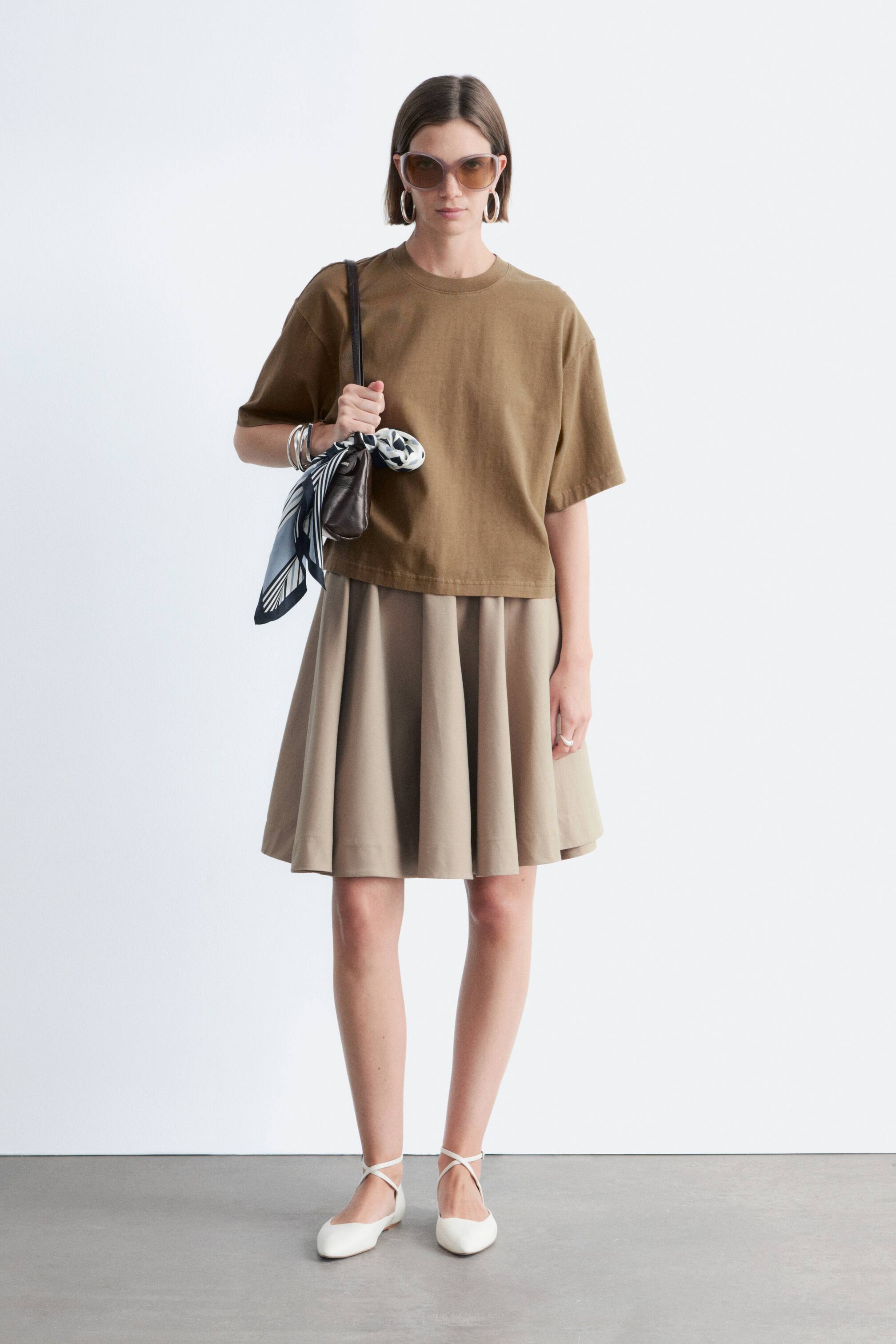 Visualizza immagine più grande: Una maglietta marrone con una forma rilassata e squadrata e maniche corte e larghe è indossata dalla modella. Presenta un classico scollo a girocollo ed è abbinata a una gonna plissettata beige.