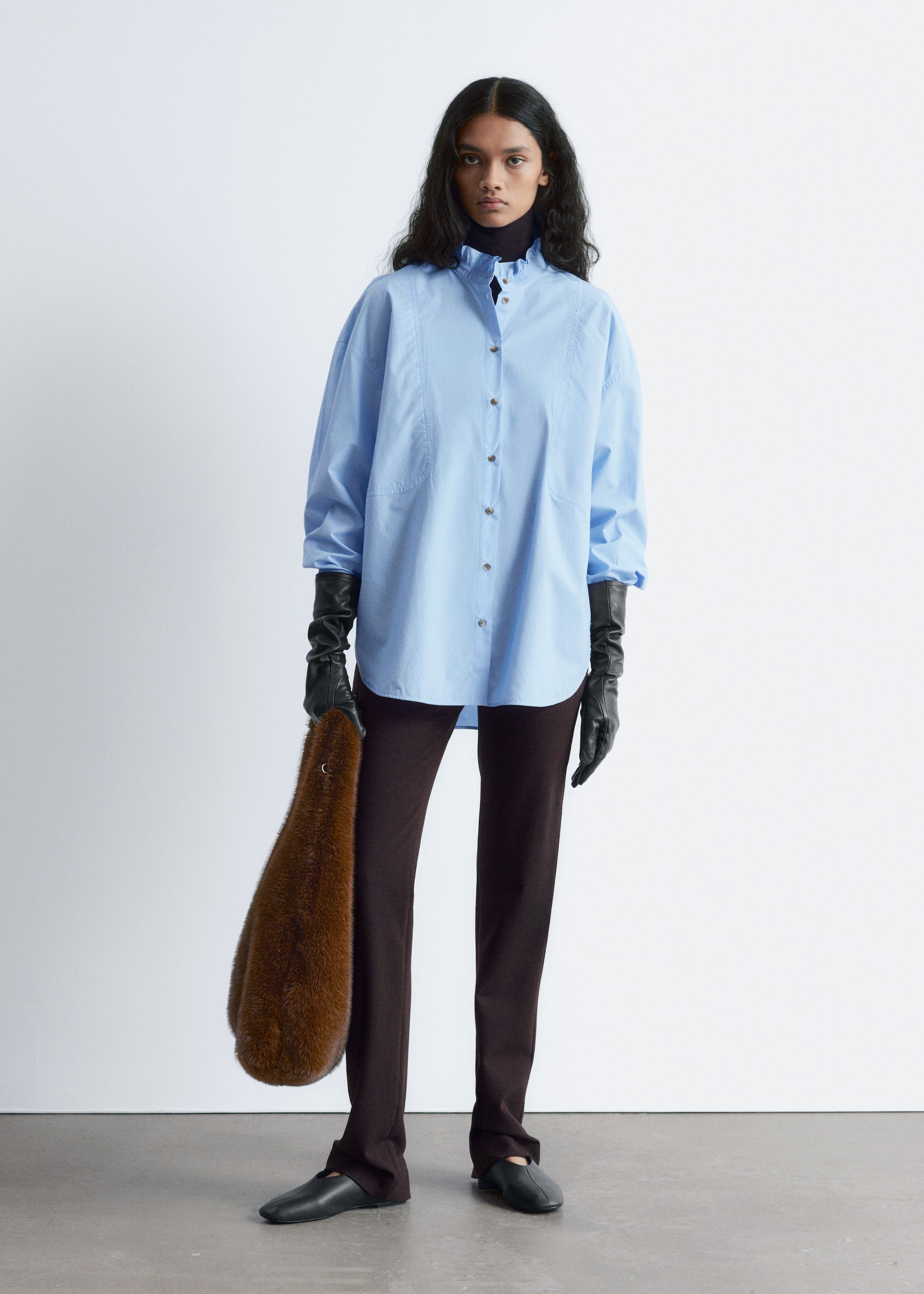 Visualizza immagine più grande: Una camicia oversize azzurro chiaro viene mostrata, caratterizzata da un colletto alto con volant e una chiusura a bottoni con bottoni scuri. Le maniche lunghe e ampie della camicia si estendono dalle spalle cadenti, indossate sotto lunghi guanti neri. La parte anteriore presenta dettagli di cuciture verticali sottili e due piccole tasche arrotondate, mentre è stratificata su un dolcevita nero.