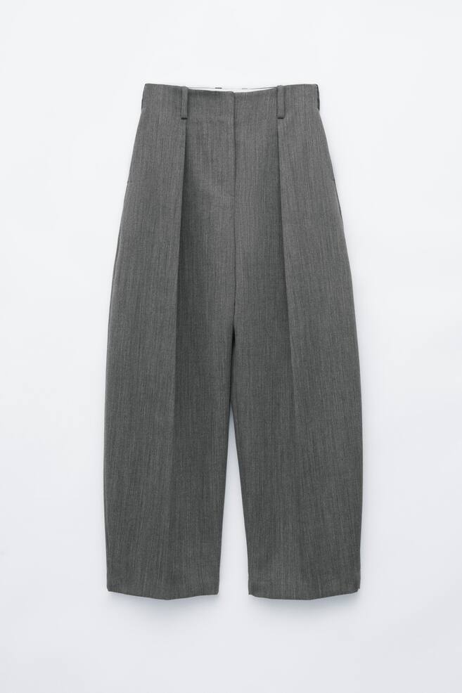 Unos pantalones grises de pernera ancha cuelgan de frente, mostrando una silueta de talle alto con pliegues frontales profundos. Los pantalones presentan una cinturilla estructurada con trabillas para cinturÃ³n y un corte de pierna relajado, ligeramente afilado. - 1