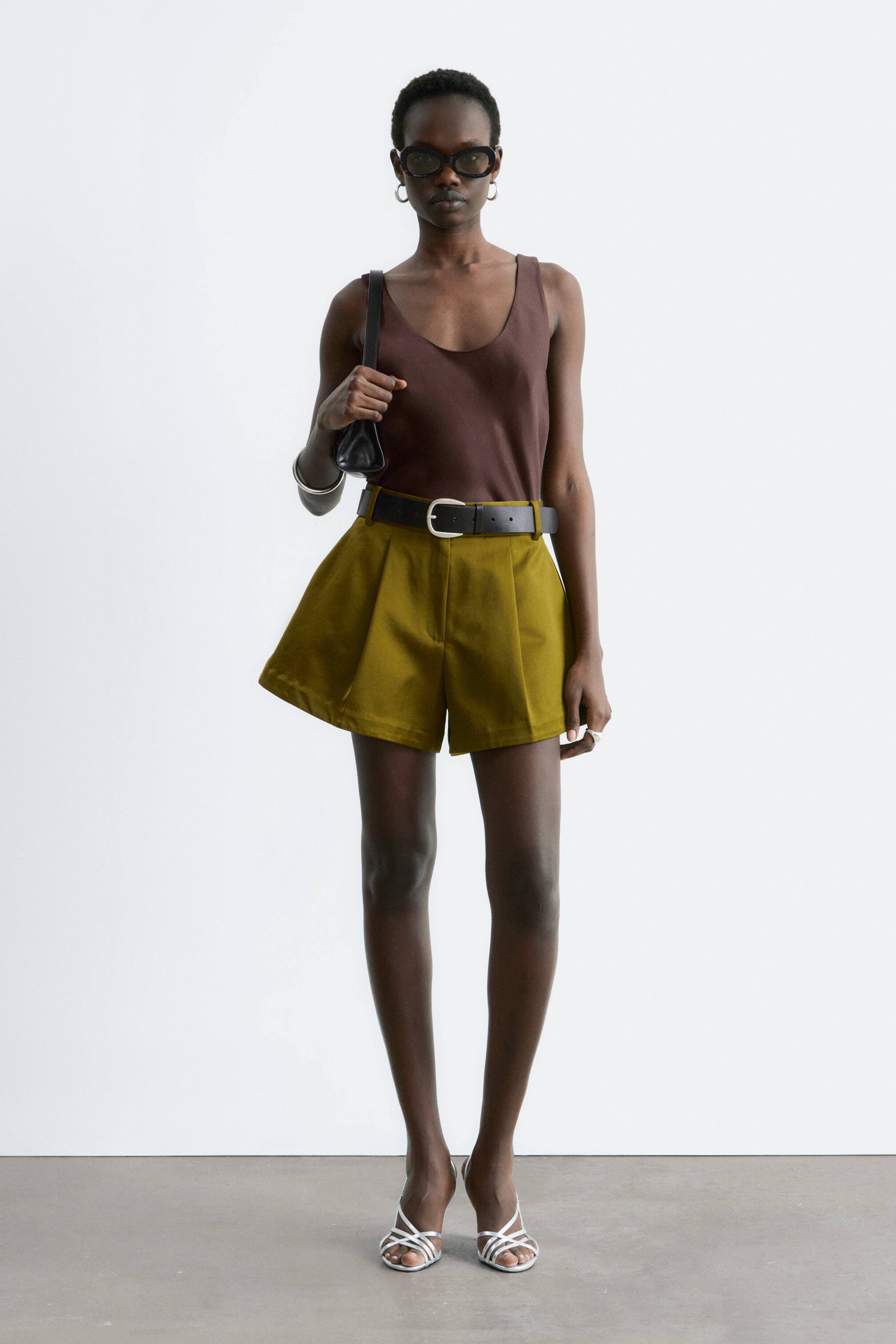 View larger image: Olive green pleated shorts feature a high-waisted fit and a flared silhouette, cinched at the waist with a black belt and a silver buckle.