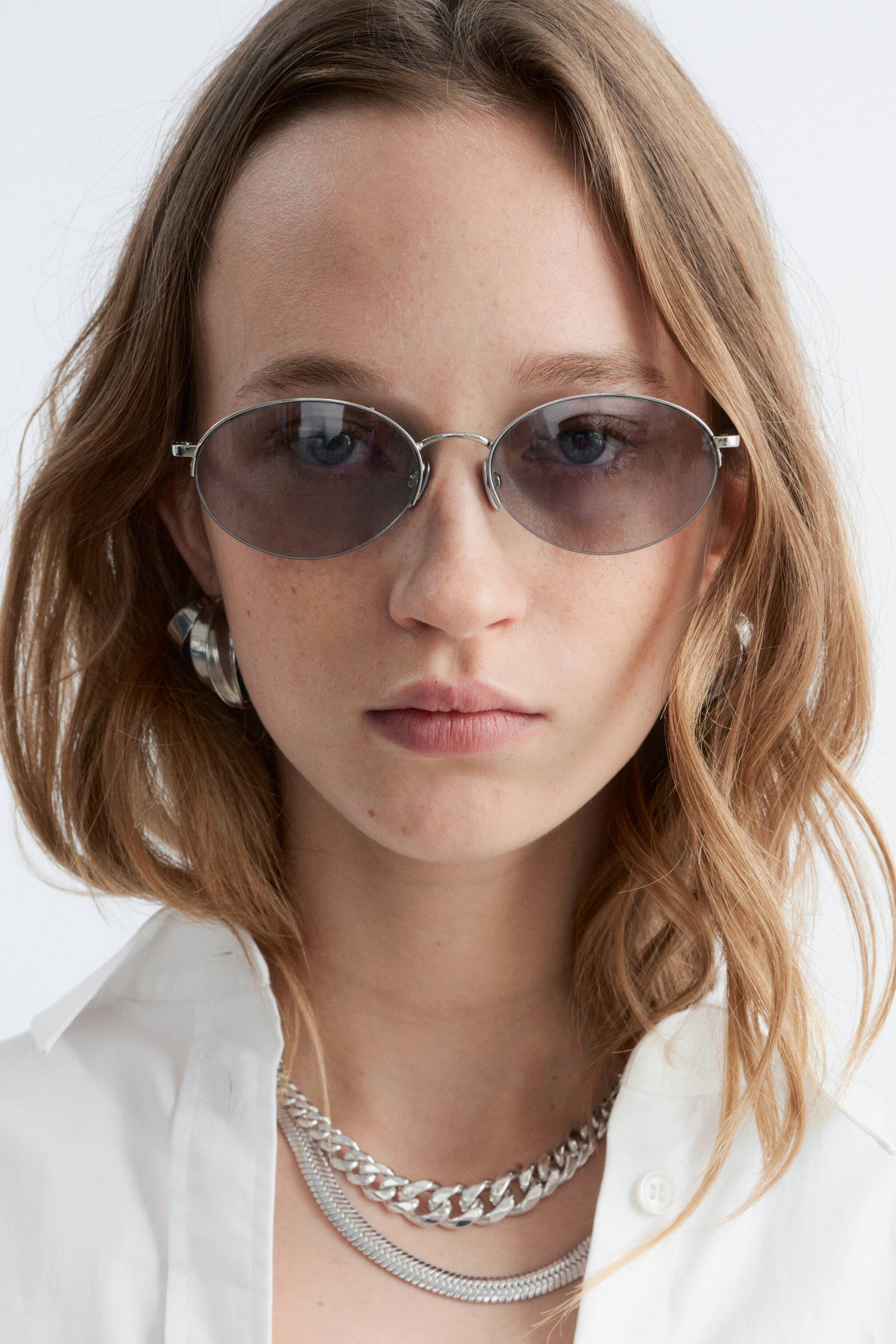 Ver imagen más grande: Las gafas de sol ovaladas presentan monturas metálicas de tono plateado delgadas y lentes tintadas en gris oscuro. Están siendo llevadas en un rostro, mirando directamente hacia adelante.