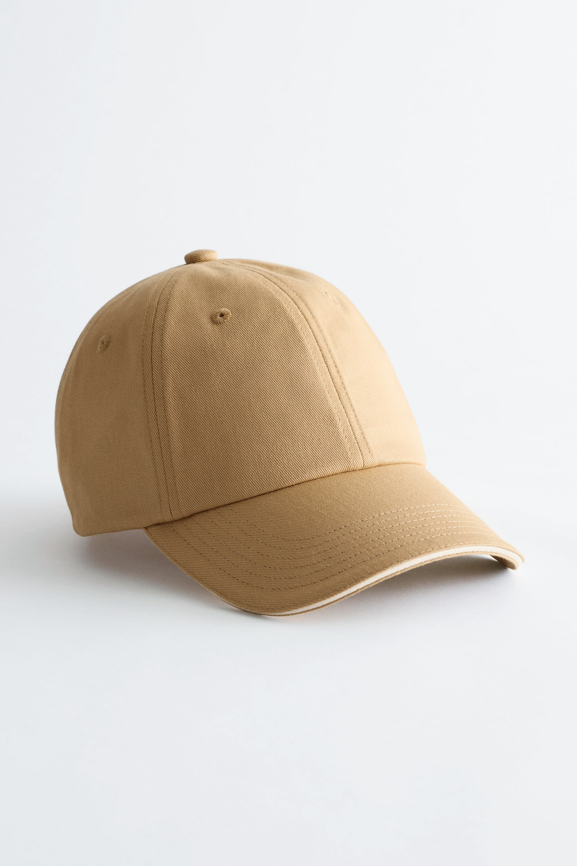 Visa större bild: En beige bomullstwillkeps presenteras från framhöger, med en paneldesign och synliga toppsömmar. Den visar en klassisk böjd skärm med flera rader av toppsömmar och ventilationshål på sidopanelerna.