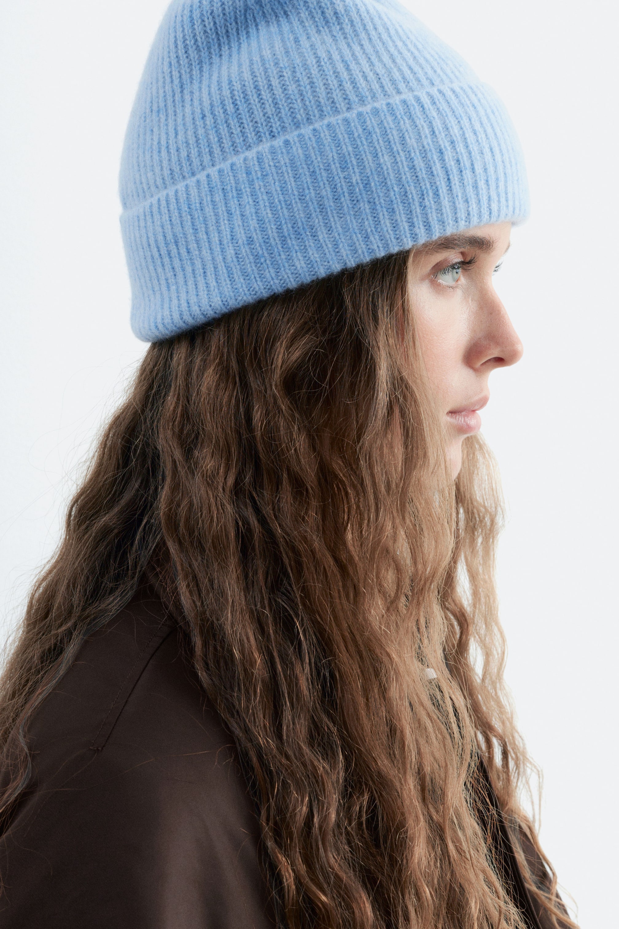Ver imagen más grande: La modelo lleva un gorro de punto acanalado de color azul claro con vuelta en el dobladillo. Se la muestra de perfil con el pelo largo y ondulado de color castaño, y lleva una chaqueta marrón oscuro.