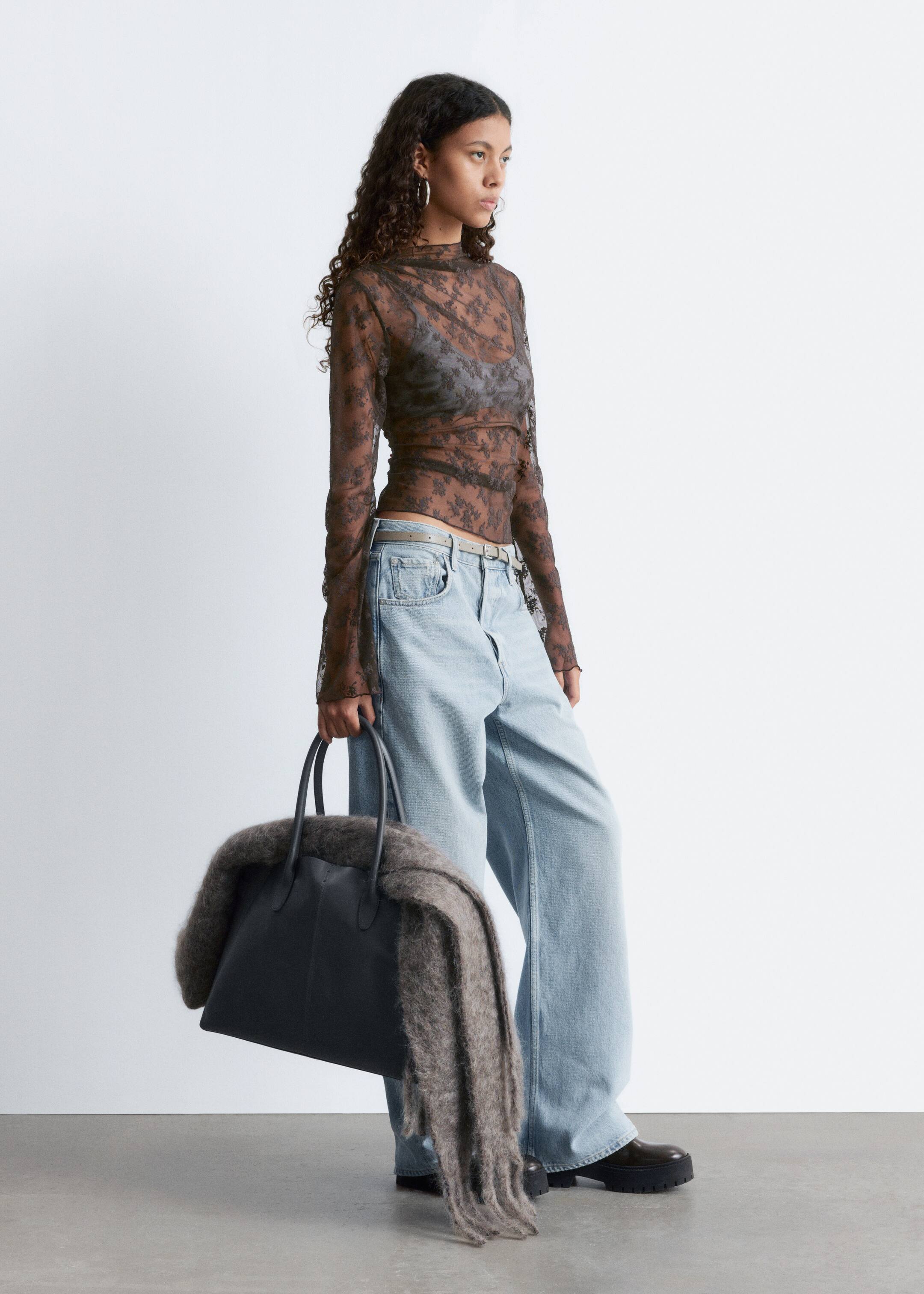 Agrandir l'image: Un haut en dentelle transparente avec un motif floral sombre et un col montant est porté par le mannequin. Le haut ajusté est doté de manches longues évasées et de fronces aux épaules, superposé sur un soutien-gorge foncé et assorti à un jean clair.