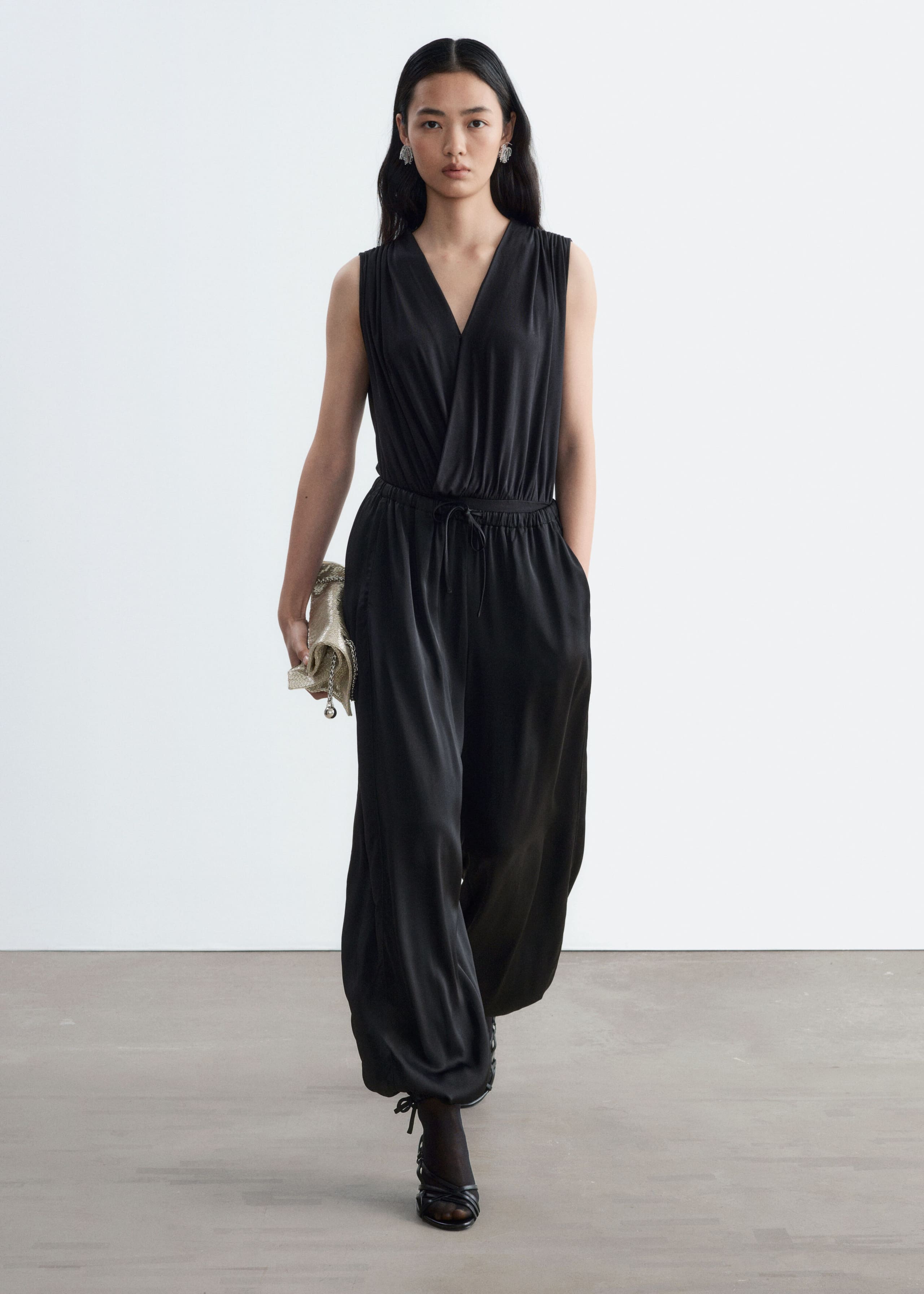 Loose-fit black trousers in a fluid, subtly shiny fabric are worn by the model. The trousers feature a balloon-leg silhouette with ties at the ankle hems and an elasticated drawstring waist, styled with a sleeveless V-neck top.
