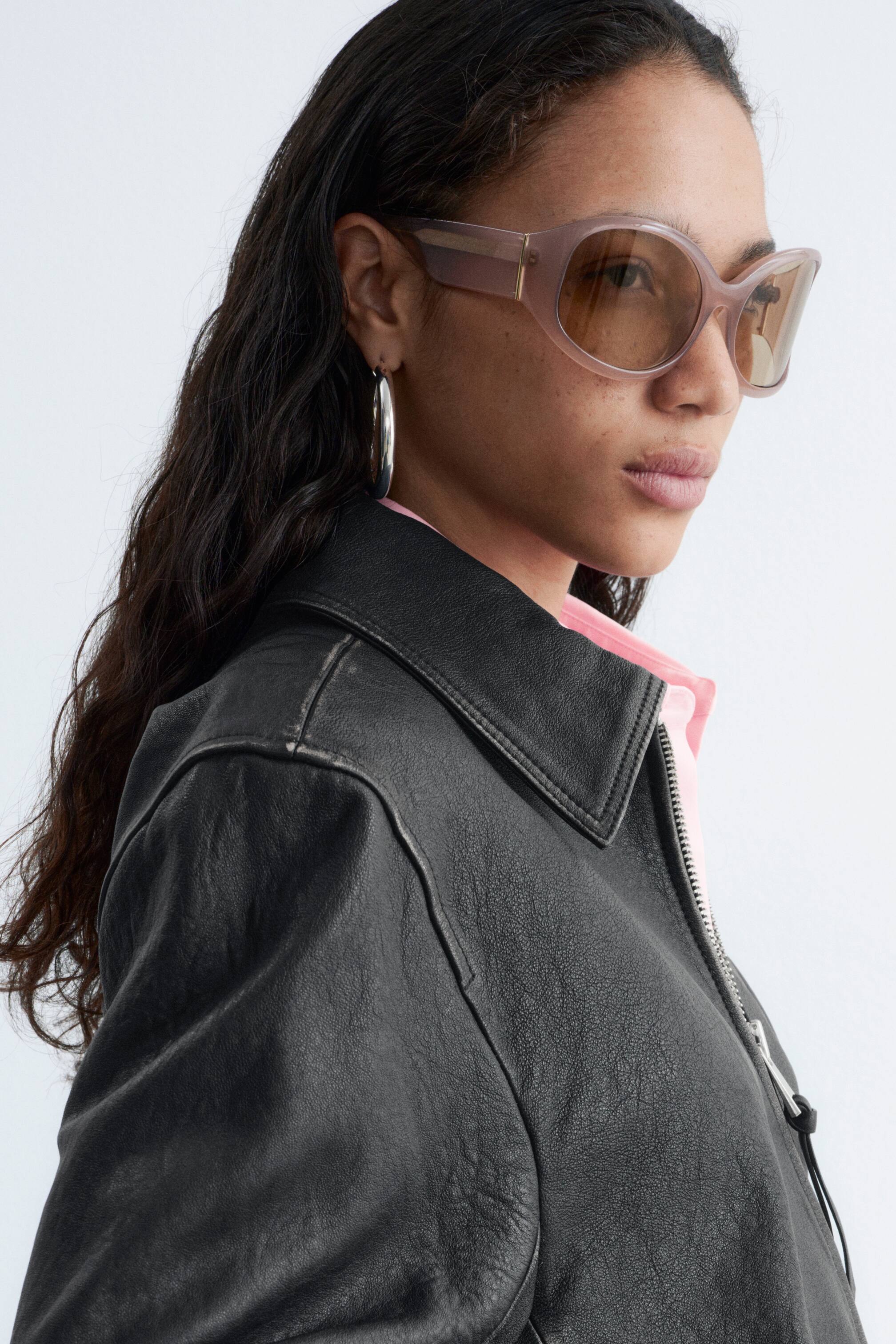 Ingrandisci l'immagine: Gli occhiali da sole presentano montature oversize, leggermente a forma di occhio di gatto, in un marrone chiaro traslucido, con lenti sfumate marroni. Mostrano un sottile dettaglio a barra color oro sulle aste, con le montature leggermente inclinate di lato.