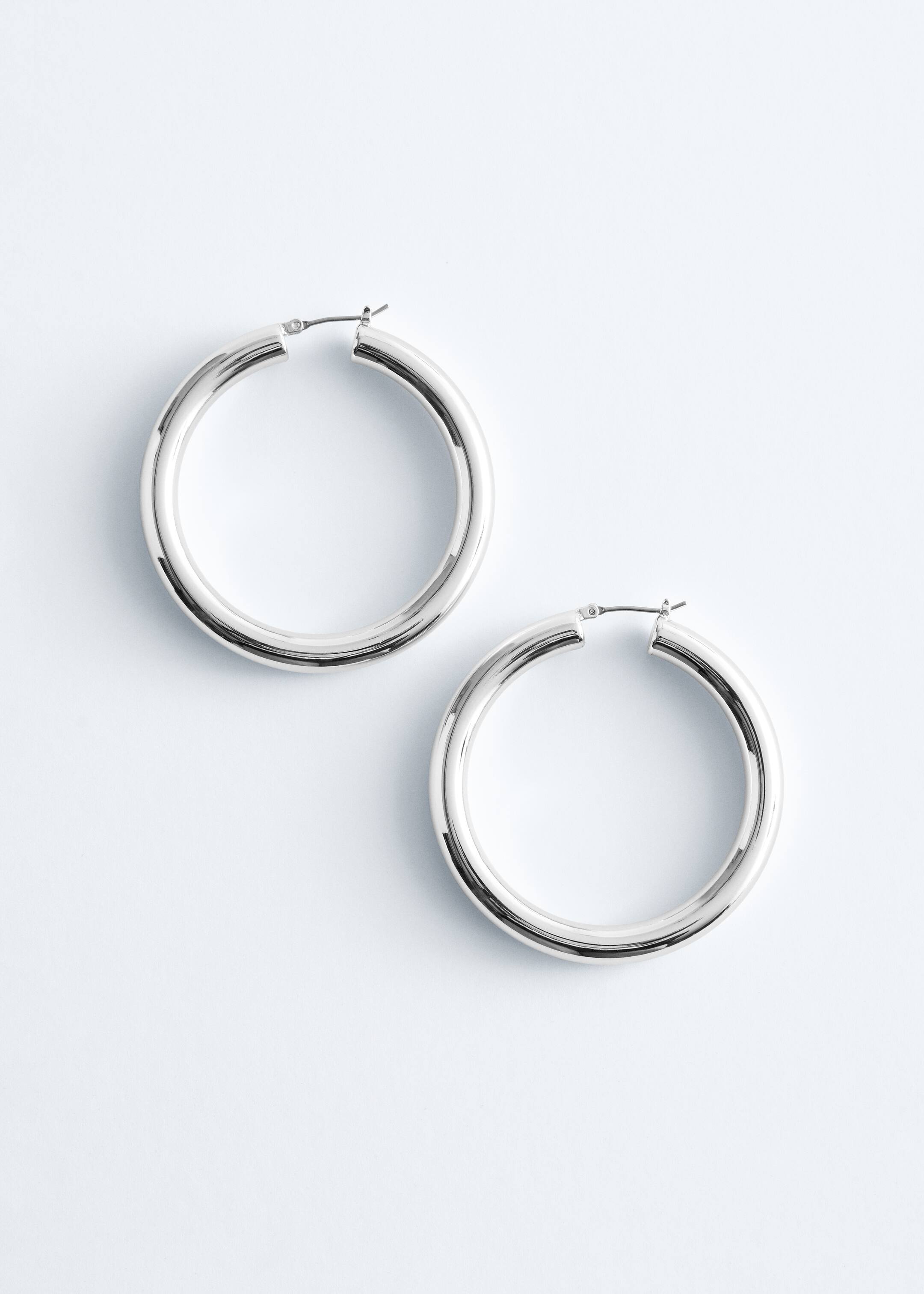 Visa större bild: Två silverfärgade hoop-örhängen ligger platt, vardera med en tjock, rörformad design och en slät, reflekterande yta. De visar sina gångjärns- och stängselsstängningar upptill och presenterar sin fulla cirkulära form.