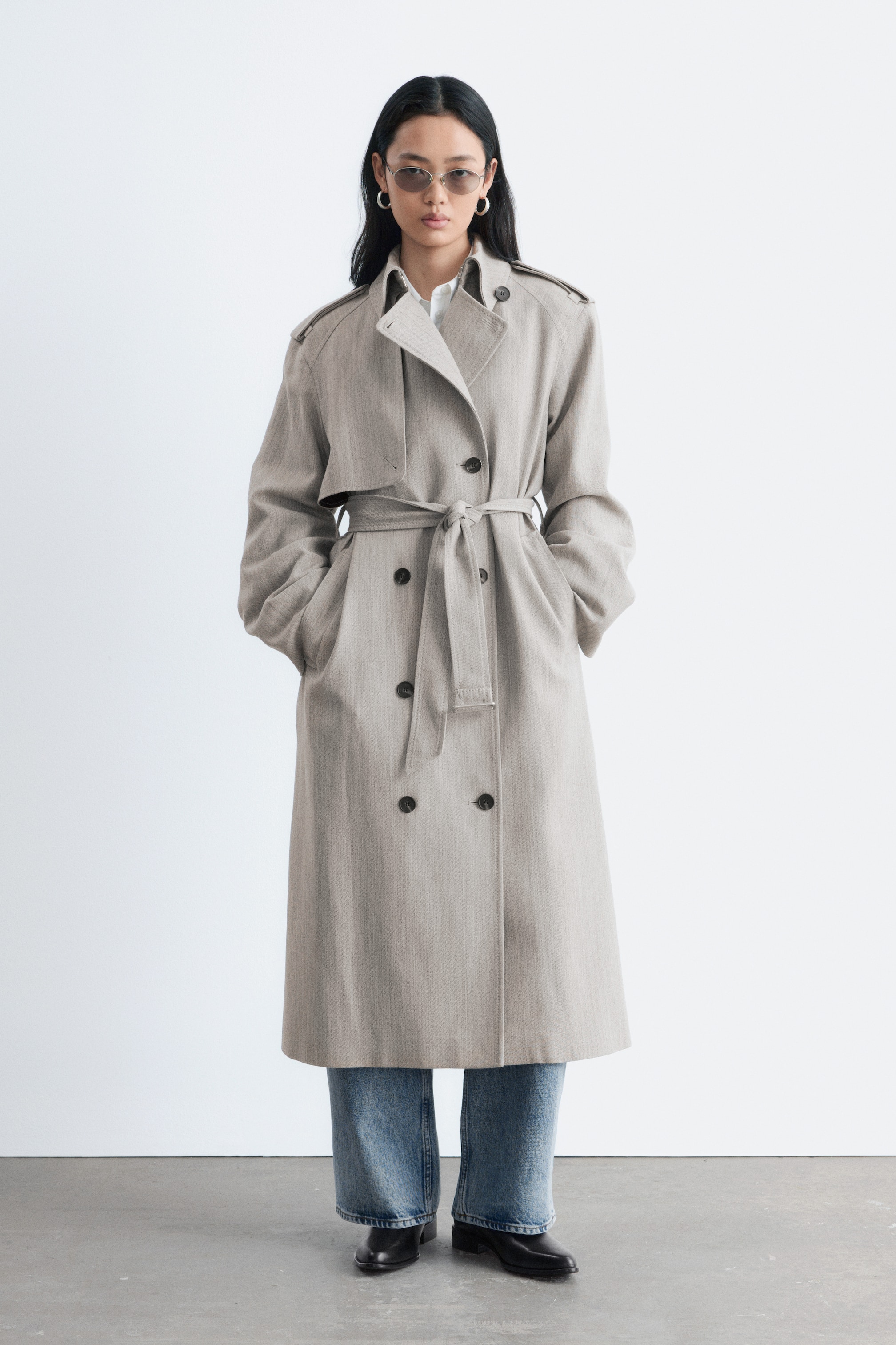 Visa större bild: En ljusbeige dubbelknäppt trenchcoat i fiskbensmönster har axelklappar, en stormklaff på höger sida och mörka knappar. Ett matchande tygbälte är knutet i midjan, och den långa kappan faller över blå jeans, med en vit skjortkrage synlig undertill.