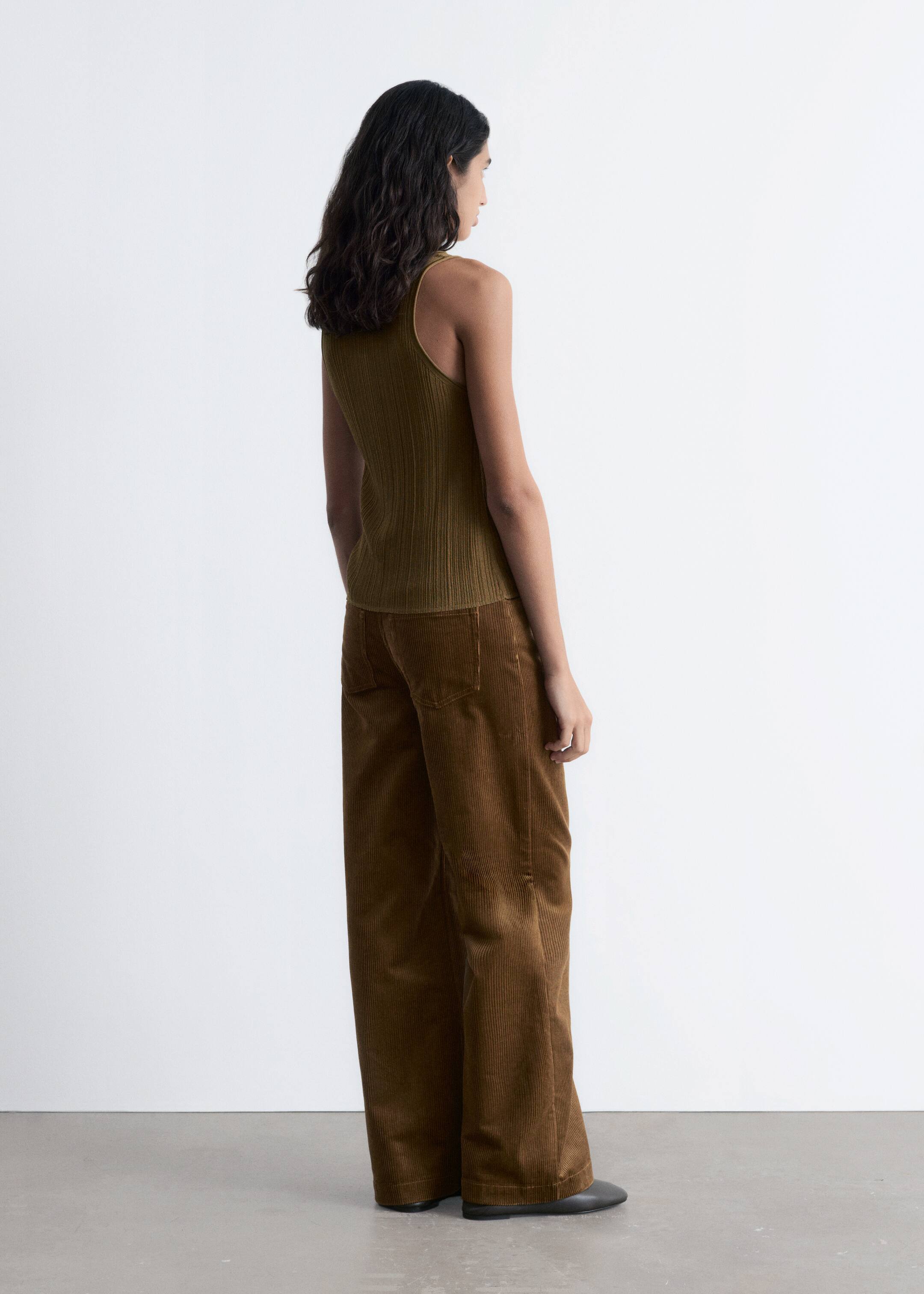 Visualizza immagine più grande: Un top a canotta in maglia a coste verde oliva è indossato, caratterizzato da una silhouette aderente e da uno scollo alto sulla schiena, visibile da un'angolazione laterale-posteriore. È abbinato a pantaloni a gamba larga in velluto a coste marroni, che mostrano la loro vestibilità rilassata e la lunghezza completa dalla stessa angolazione.