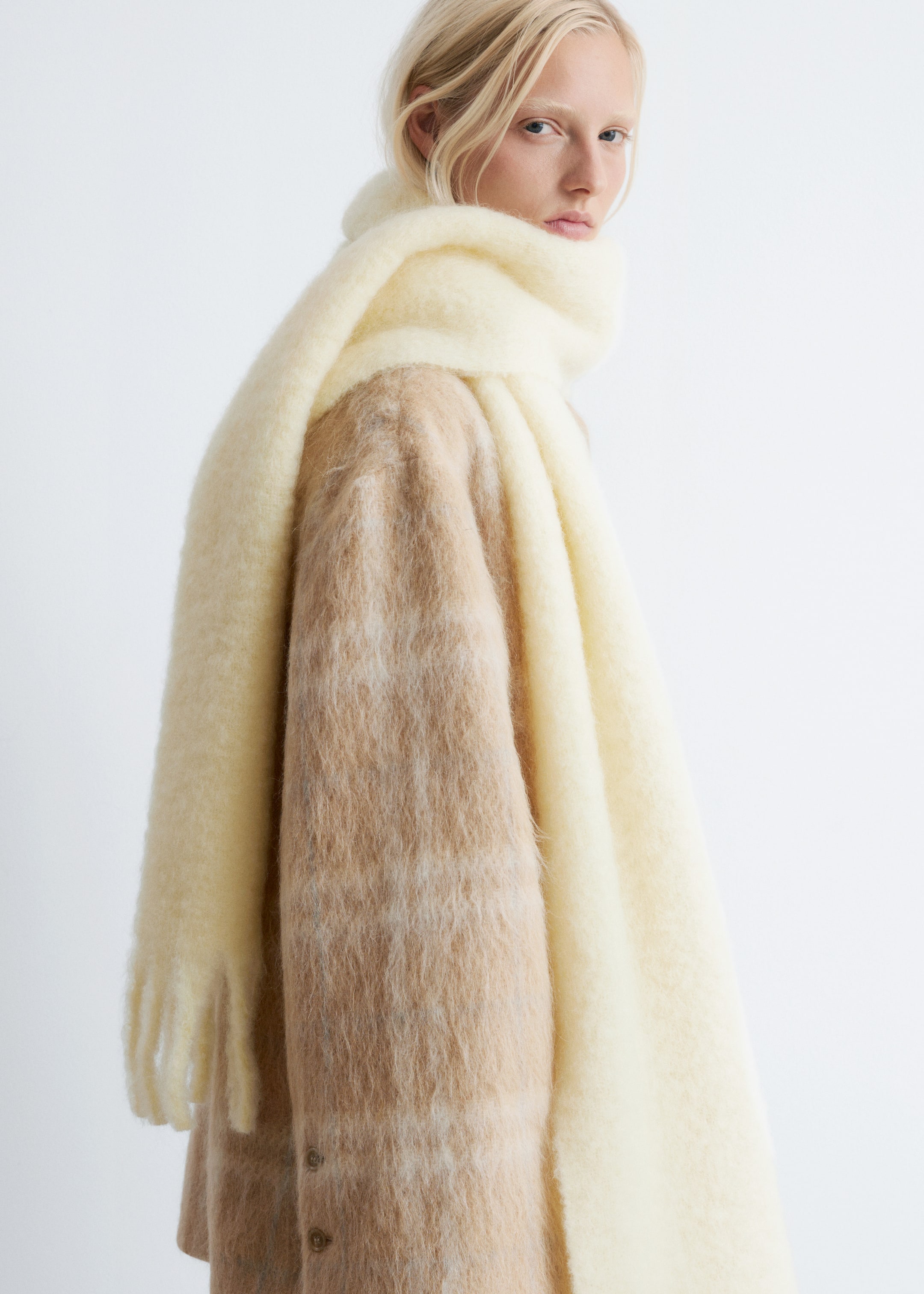 Visualizza immagine più grande: Maxi sciarpa in misto mohair di colore crema chiaro, caratterizzata da una texture pelosa e frange alle estremità, indossata dalla modella avvolta intorno al collo e alle spalle sopra un cappotto a fantasia e strutturato.