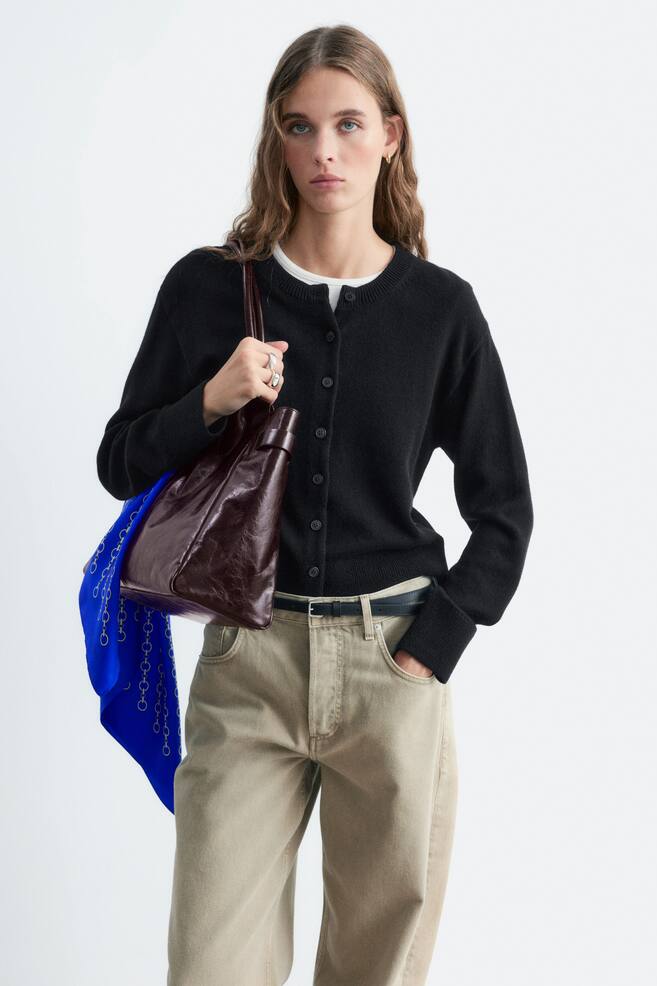 Een zwart vest met ronde hals en knoopsluiting aan de voorkant wordt door het model gedragen over een wit T-shirt. Het vest is gestyled met lichtbeige broek en een glanzende, donkerbruine leren schoudertas die door het model wordt vastgehouden. - 1