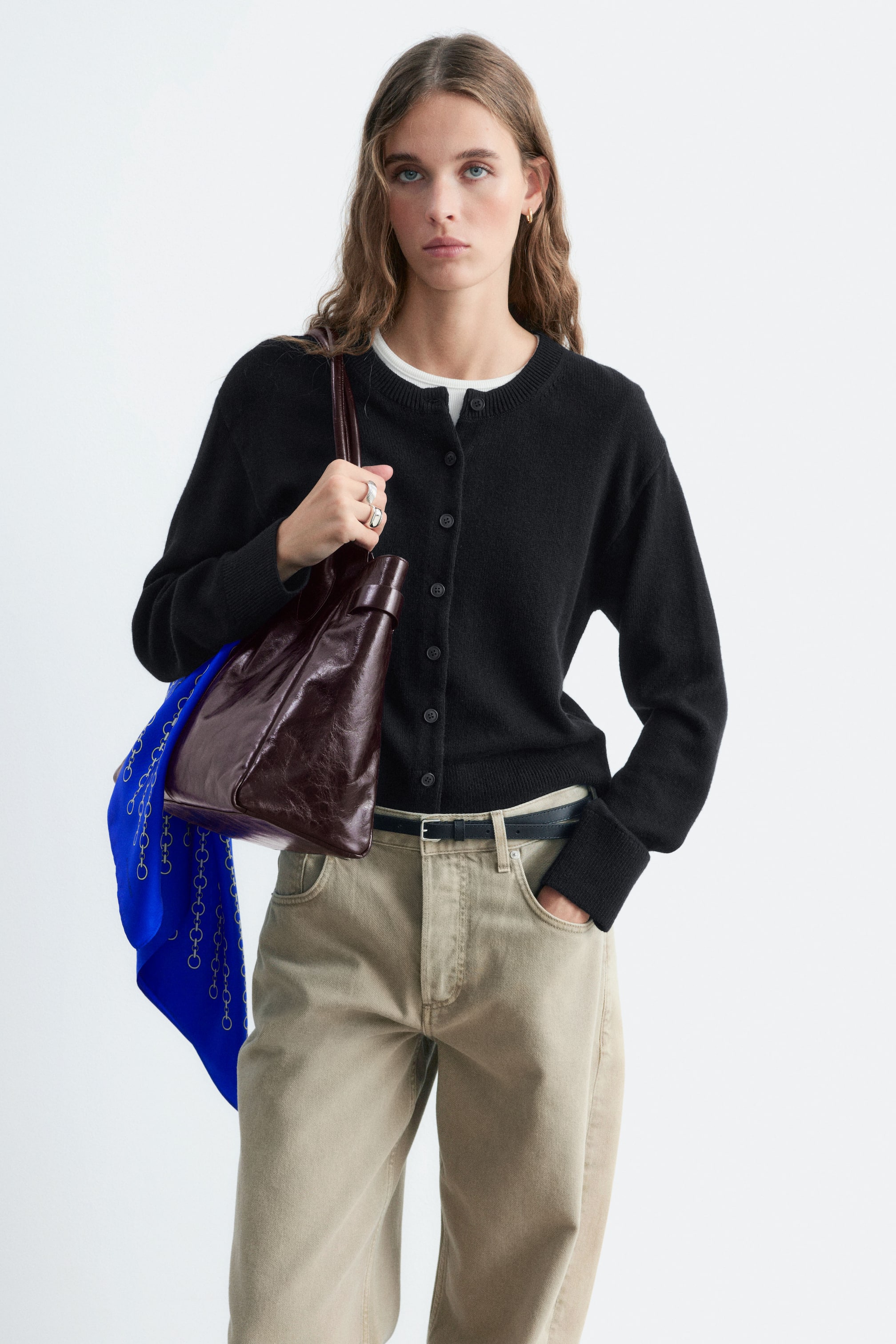 Se større billede: En sort cardigan med rund hals og knaplukning foran bæres af modellen over en hvid T-shirt. Cardiganen er stylet med lysebeige bukser og en blank, mørkebrun læder skuldertaske, som modellen holder.