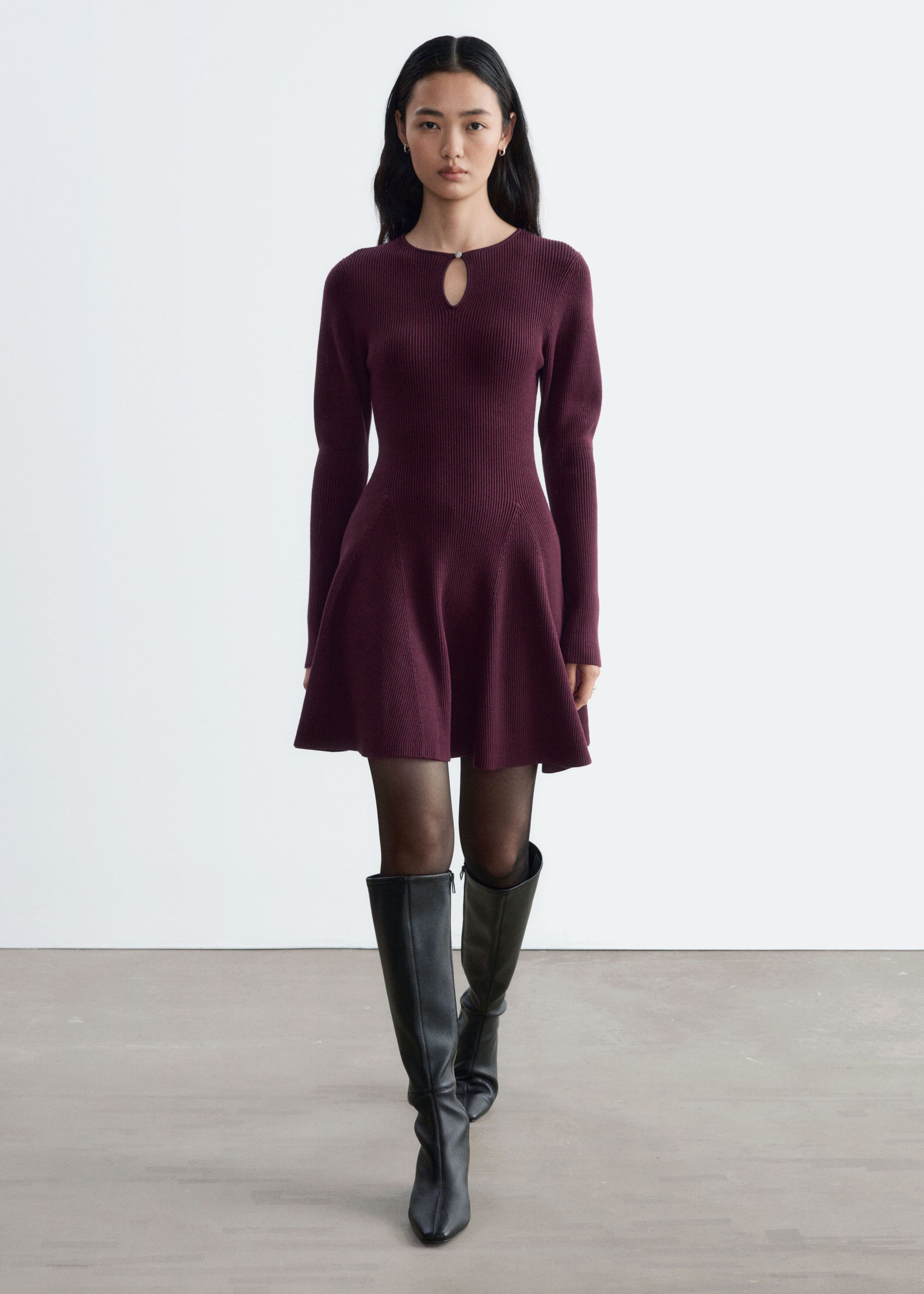 A ribbed knit mini dress in deep burgundy is being worn by the model, featuring long sleeves, a keyhole cutout at the neckline, and a flared paneled skirt. The dress is styled with black sheer tights and knee-high black leather boots.