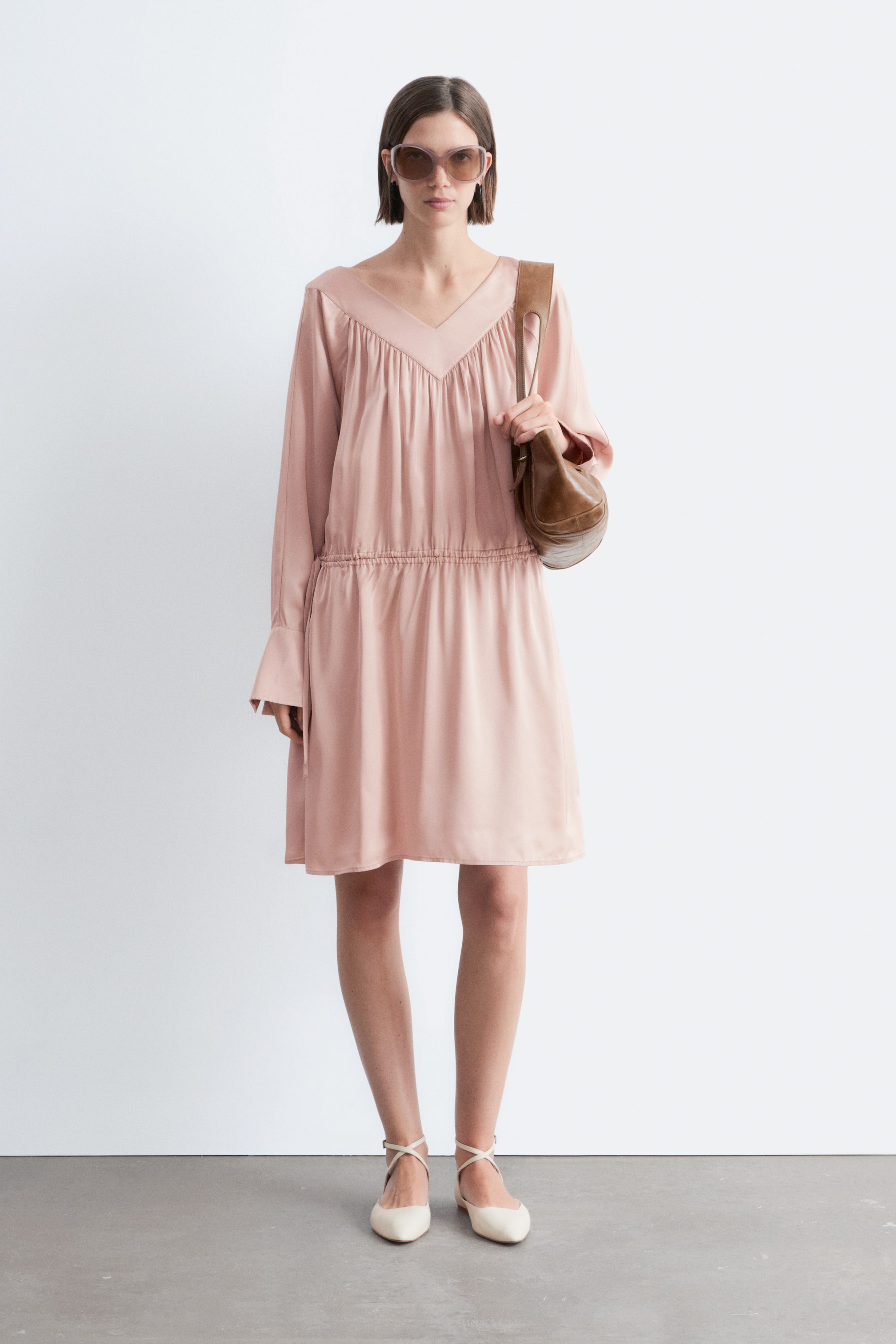 Vis større bilde: En lyserosa knelang kjole med V-hals, rynket liv og lange ermer, som fremhever en elastisk midje og et flytende skjørt. Den styles med en brun skulderveske og kremfargede kryssede ballettsko.