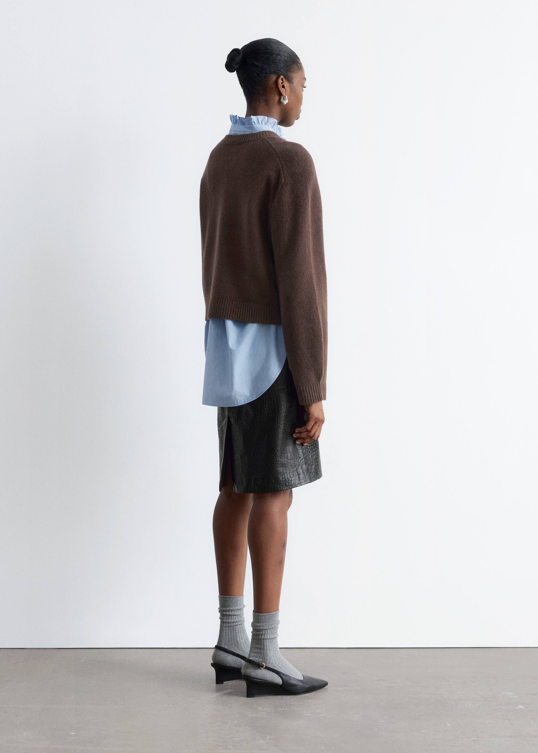 Grösseres Bild ansehen: Aus einer 3/4-Rückansicht wird ein brauner Pullover mit Raglanärmeln und geripptem Saum über ein hellblaues Hemd mit gerüschtem Kragen und abgerundetem Saum getragen. Ein schwarzer knielanger Rock mit strukturierter Oberfläche und einem Rückenschlitz wird ebenfalls getragen, zusammen mit schwarzen Slingback-Schuhen und grauen gerippten Socken.