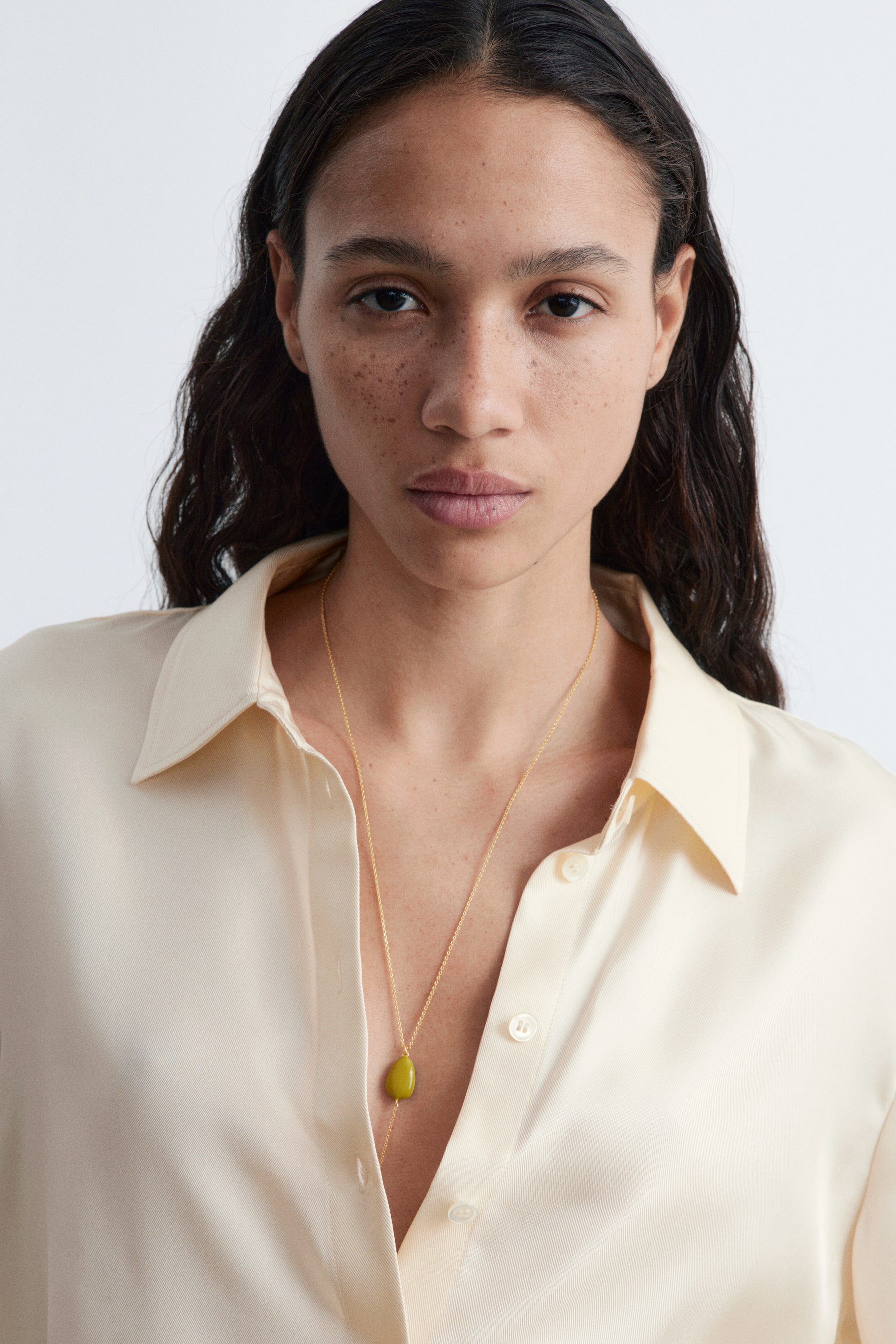 Agrandir l'image: Un collier présente une chaîne dorée avec un pendentif unique, lisse, de forme irrégulière et de couleur vert jaunâtre. Il est stylisé en lariat, avec la chaîne formant une forme en Y et le pendentif reposant sur une chemise à col ouvert de couleur crème.