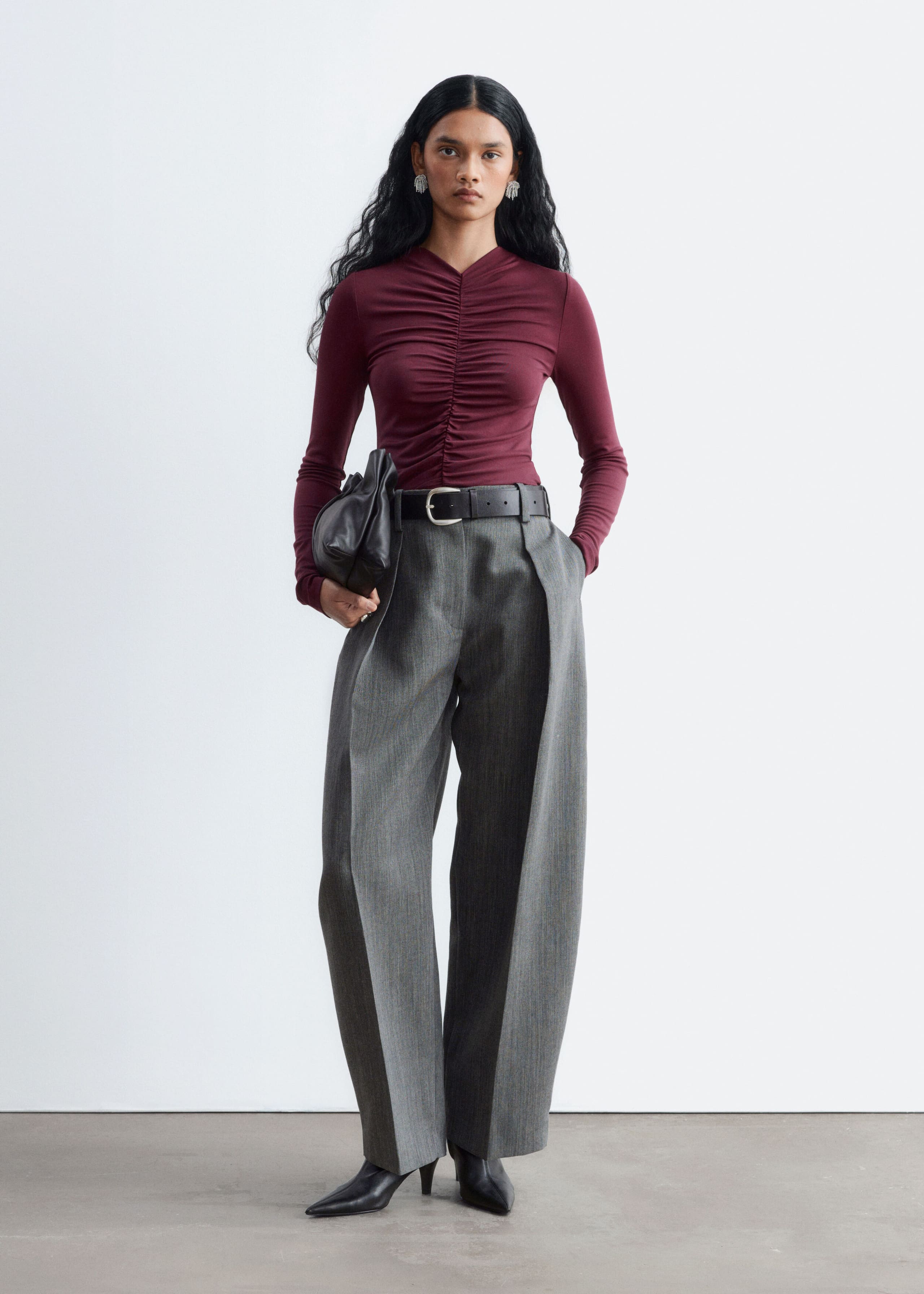A burgundy long-sleeve top with a gathered center-front seam is worn by the model, tucked into wide-leg gray trousers. The model is holding a black clutch bag under one arm, showcasing the fitted silhouette and ruched detailing of the top.