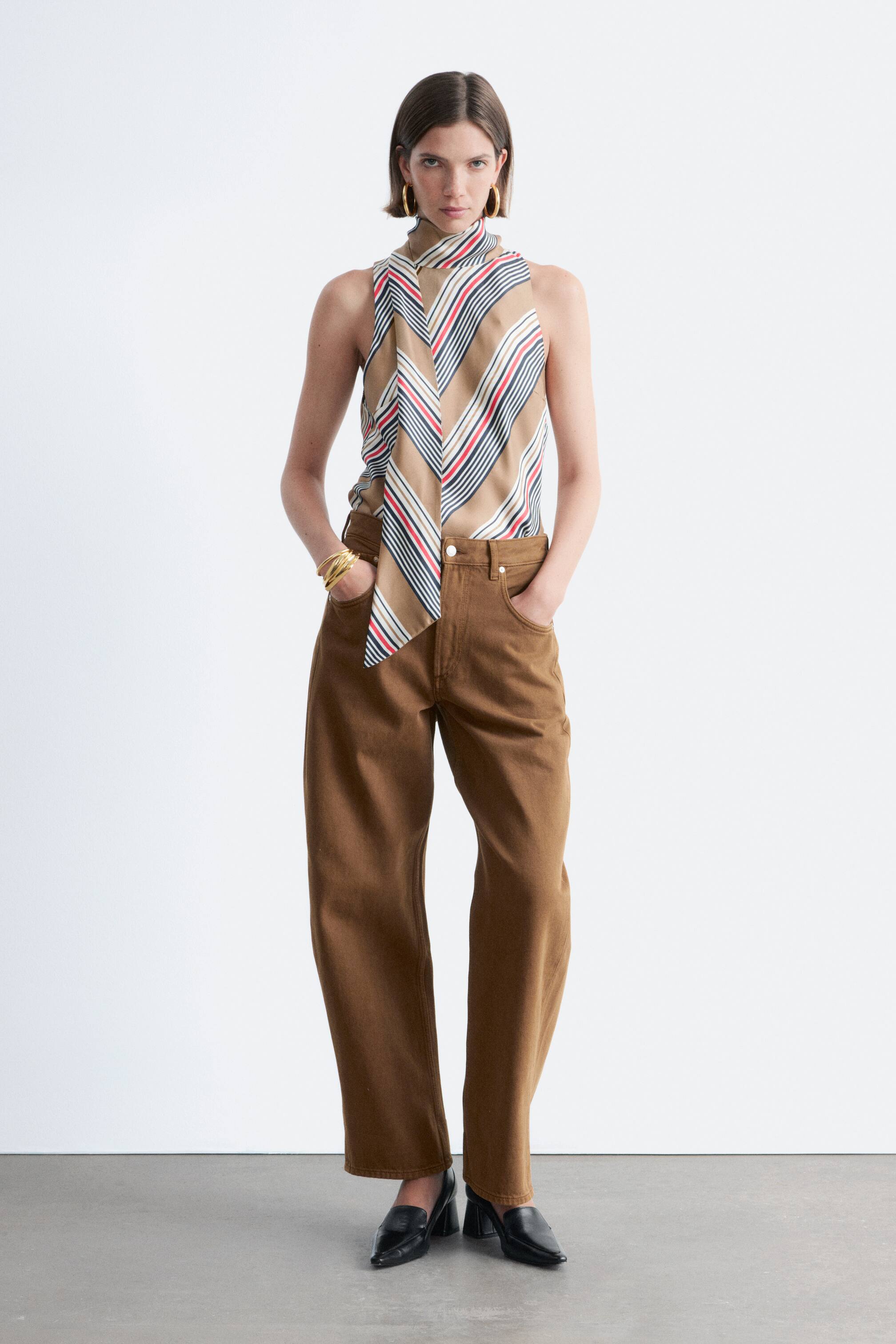 Ingrandisci l'immagine: Un outfit a figura intera viene presentato, caratterizzato da un top senza maniche con un motivo a righe diagonali nei colori beige, bianco, rosso e nero, e un collo alto con fiocco, infilato in pantaloni marroni a gamba larga con tasche frontali. Con i pantaloni vengono indossati dei sabot neri a punta con tacco a blocco.