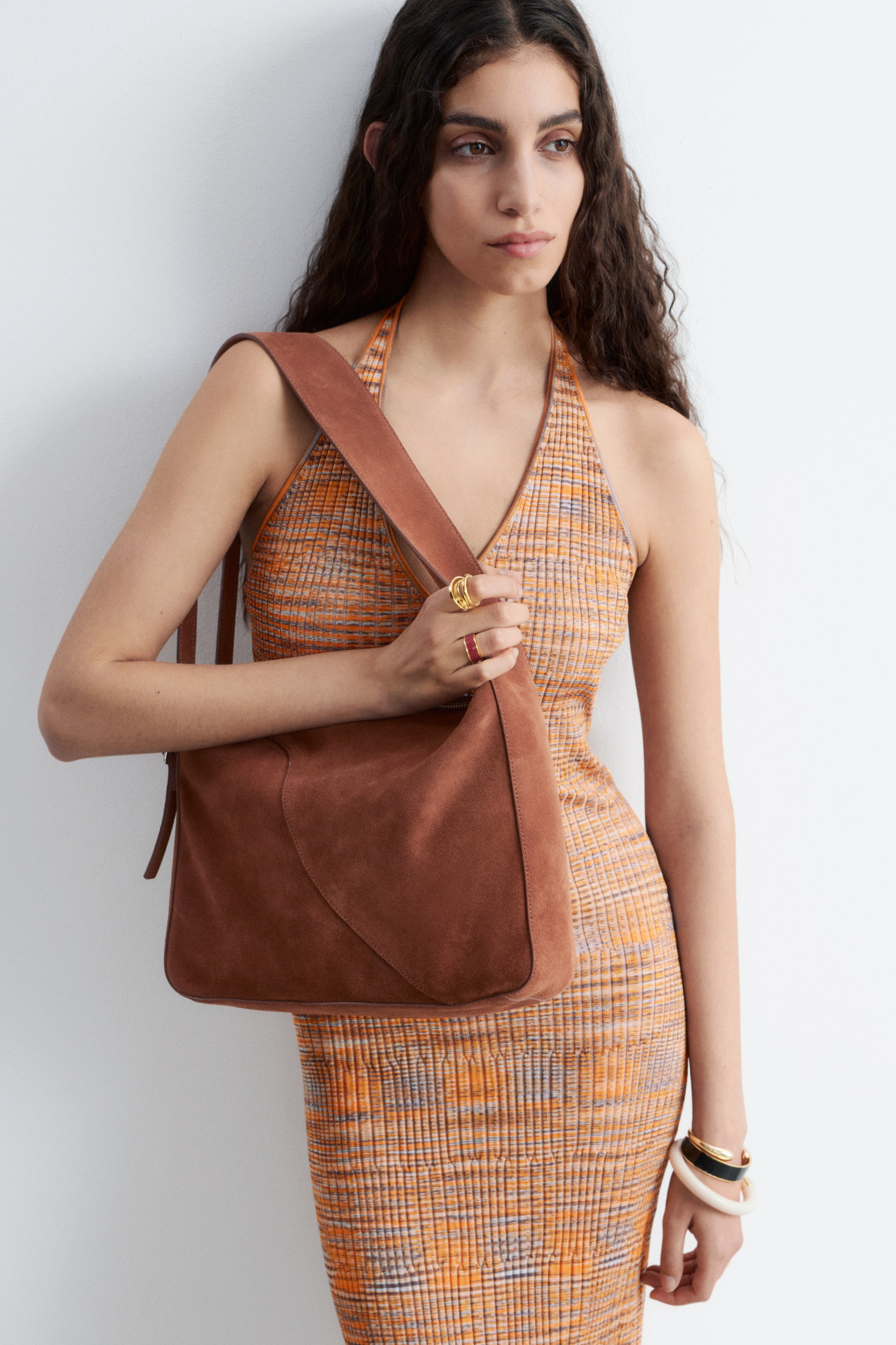 Zobraziť väčší obrázok: Hnedá semišová kabelka s čistou, zaoblenou siluetou je nesená na pravom ramene modelky cez vzorované šaty. Pravá ruka modelky spočíva na tele kabelky, ktorá má široký, plochý popruh.