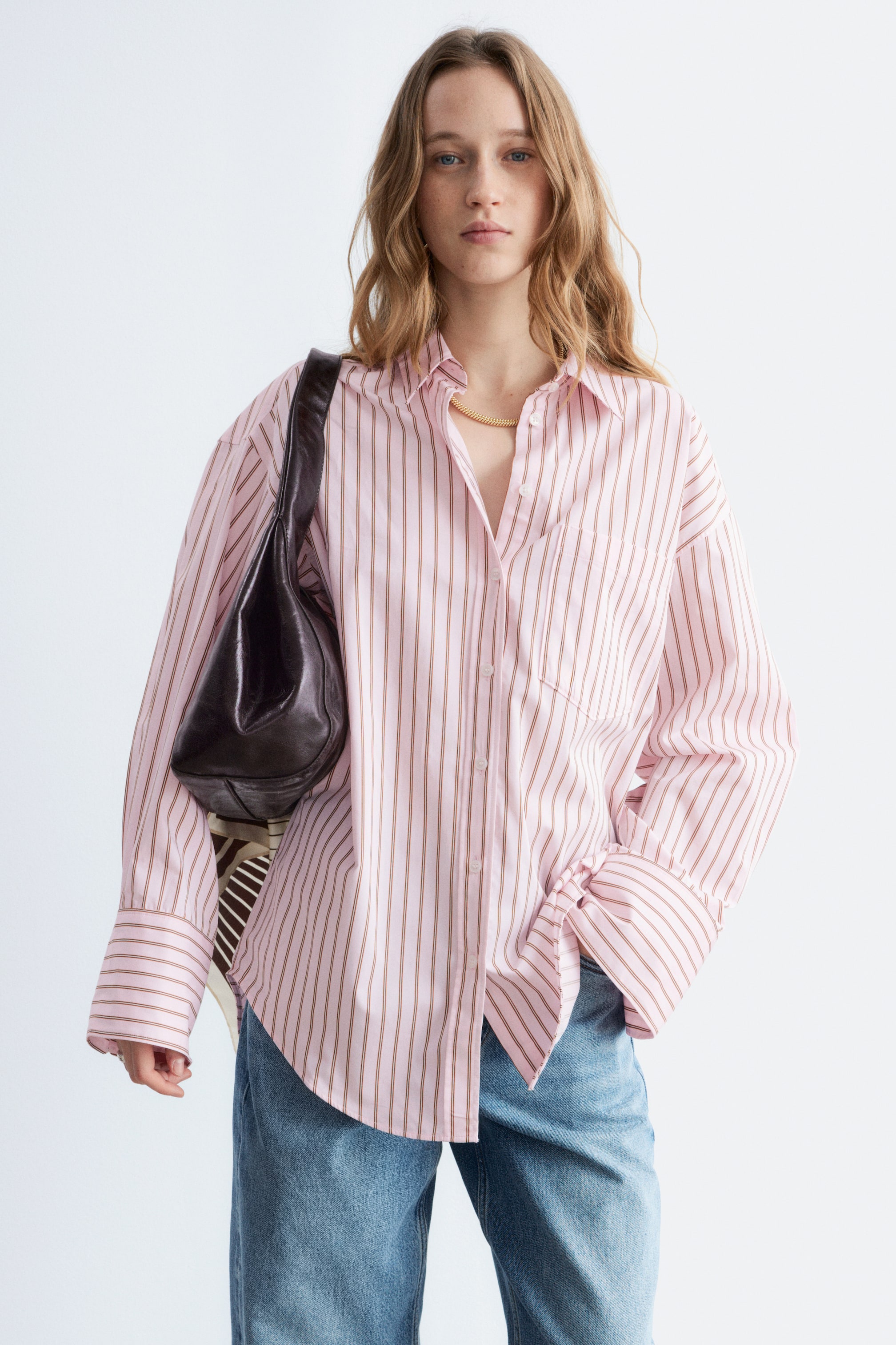 Visa större bild: Den rosa randiga skjortan har tunna bruna vertikala ränder, en spetsig krage och en bröstficka på vänster sida. Den har långa, avslappnade ärmar med breda manschetter, varav en är uppvikt, och dess främre fåll är knuten i en knut.