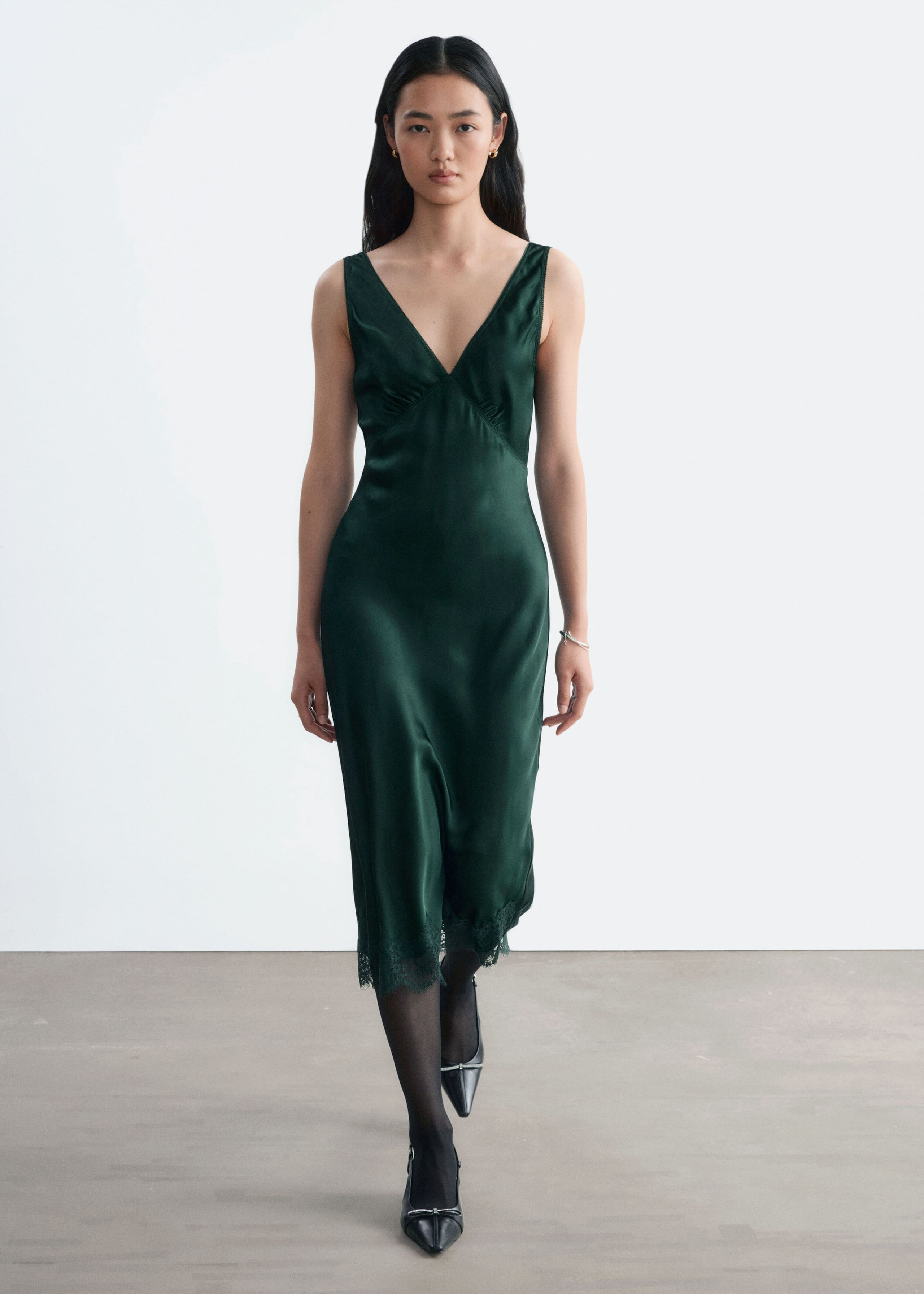 A dark green satin slip dress featuring a V-neckline, gathered bust, and delicate lace hem is worn by the model. The dress is styled with sheer black tights and black pointed-toe shoes.