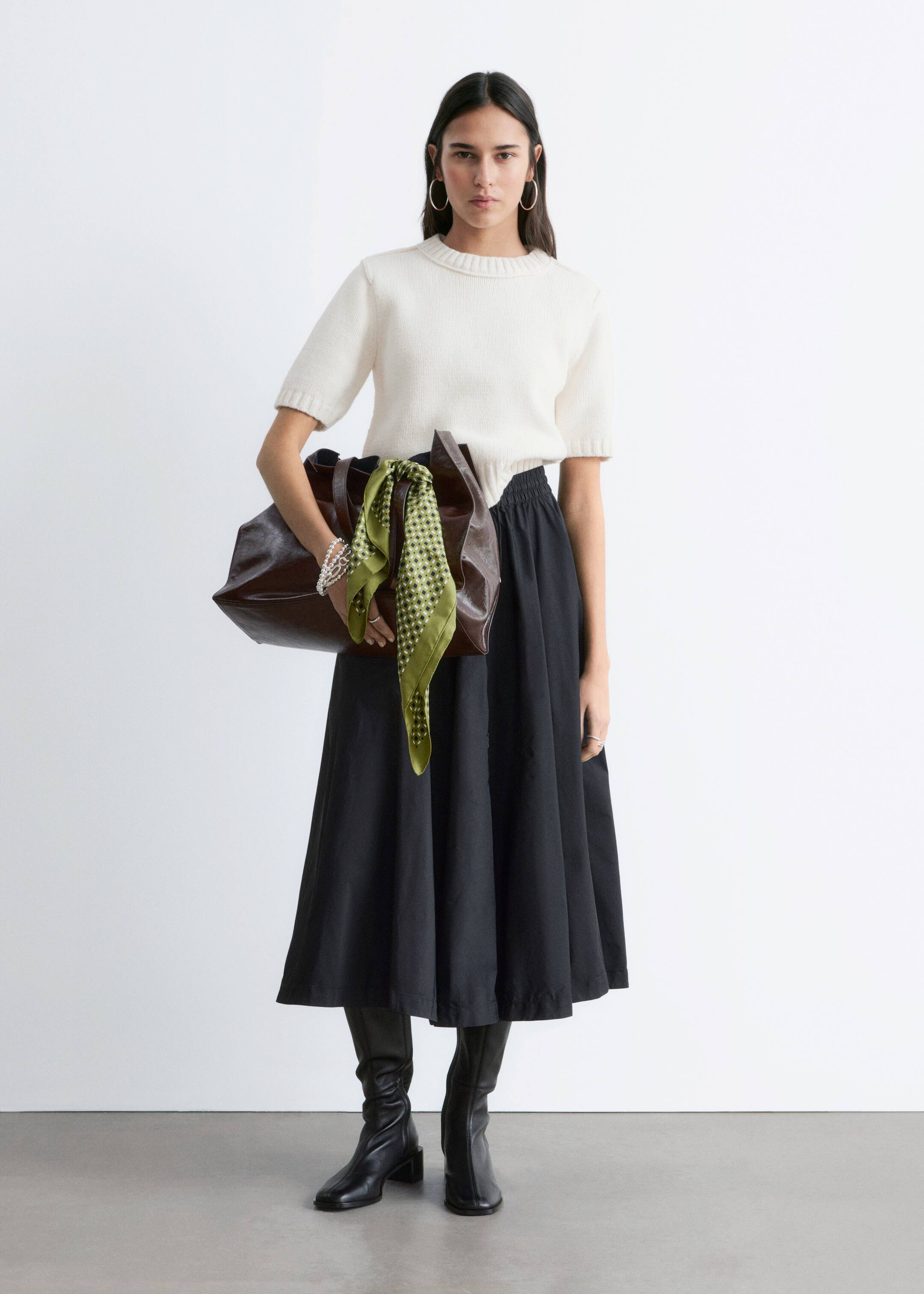 Vezi imaginea mai mare: Un top tricotat cu mâneci scurte, de culoare alb murdar, prezintă un guler rotund și manșete cu nervuri, fiind asortat cu o fustă neagră de lungime medie, care are o talie elastică adunată și o siluetă evazată în formă de A. Cizmele negre până la genunchi completează ansamblul, alături de o geantă tote din piele maro închis și o eșarfă cu model verde, care este drapată peste geantă.