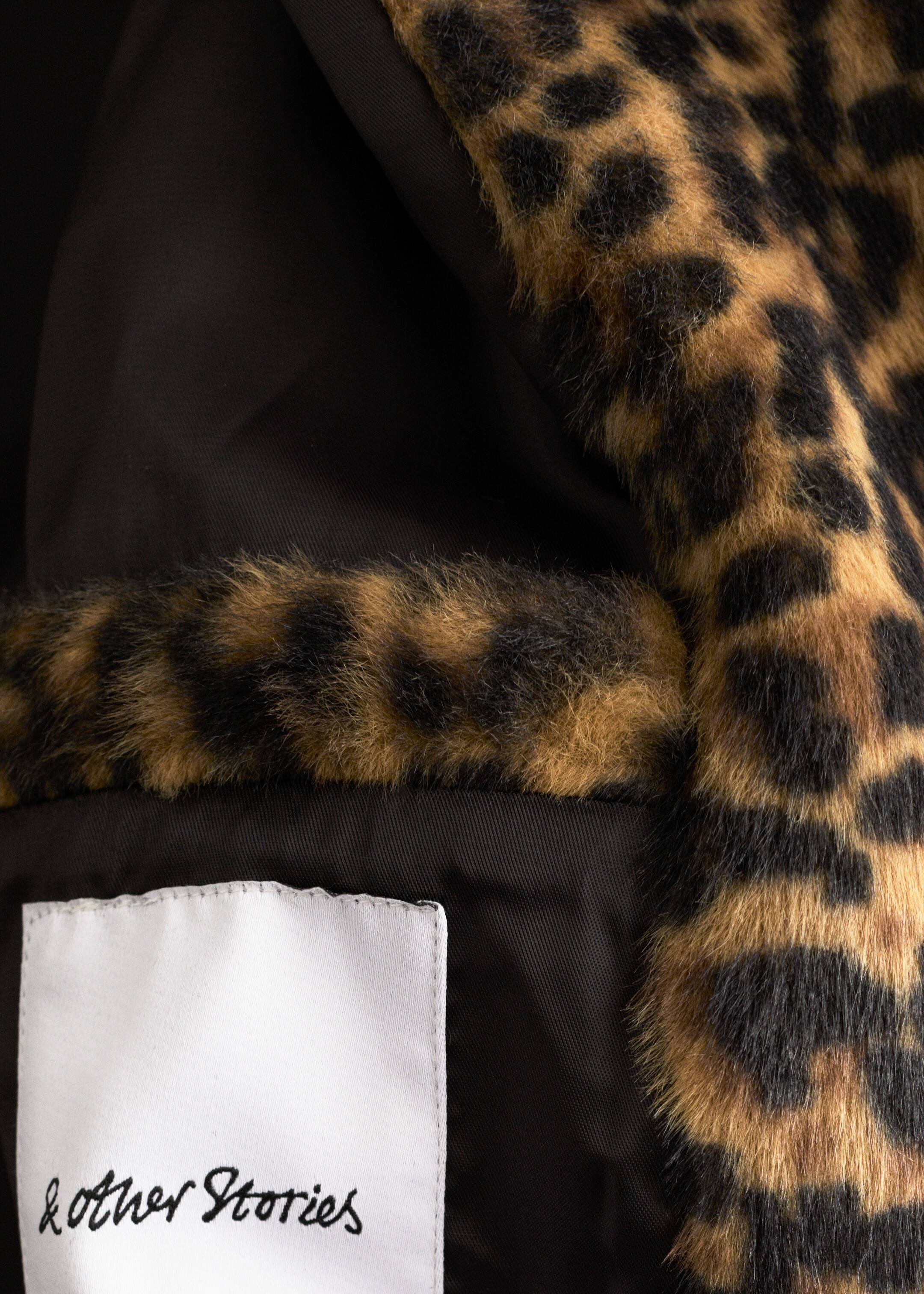 Se større billede: Indersiden af et tøjstykke viser en sort foring, der grænser op til en leopardmønstret faux fur yderside. Et hvidt stofmærke med teksten '& Other Stories' er syet på den sorte foring.
